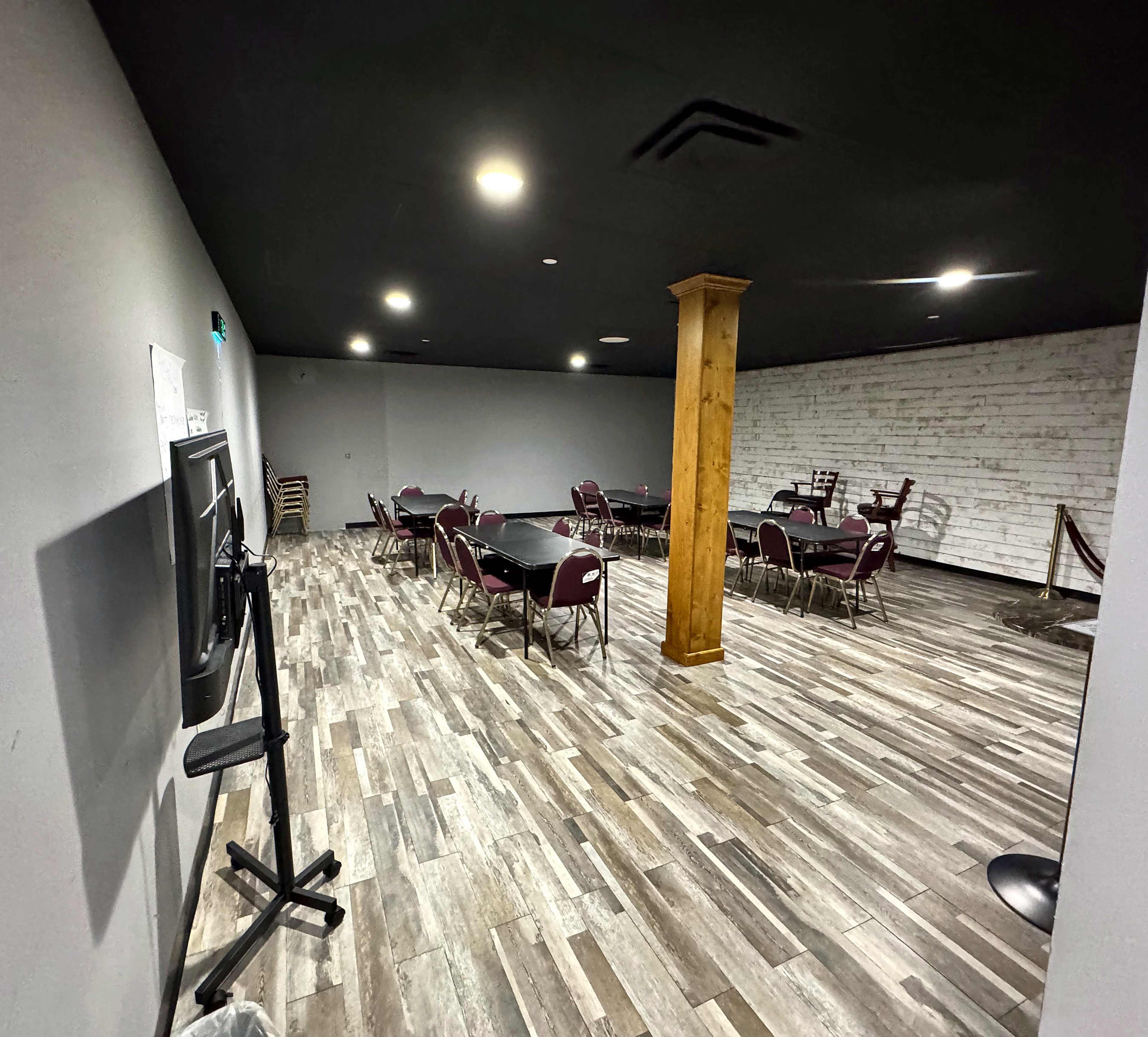 The image shows a spacious room with wooden flooring, featuring several tables and chairs set up for dining or meetings, with a column in the center and white brick walls on one side.