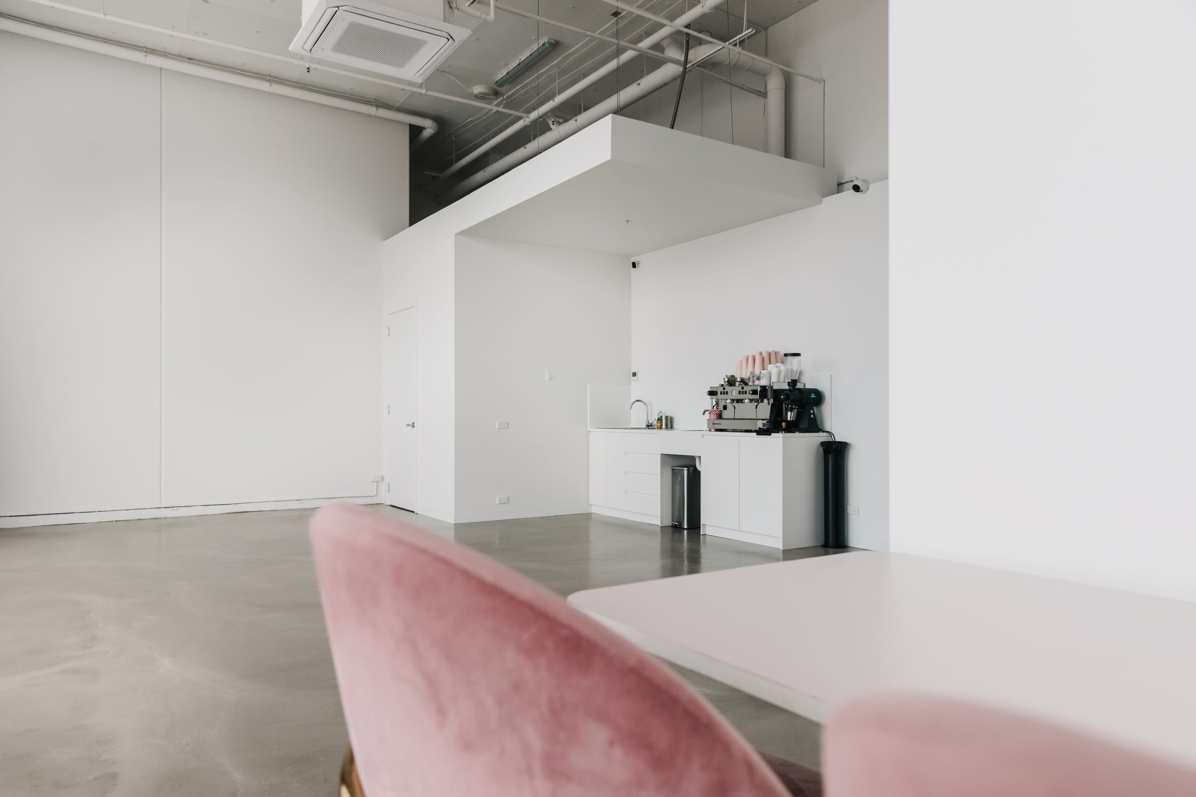 The image shows a modern interior space featuring a pink chair with a clean white wall and a coffee station in the background.