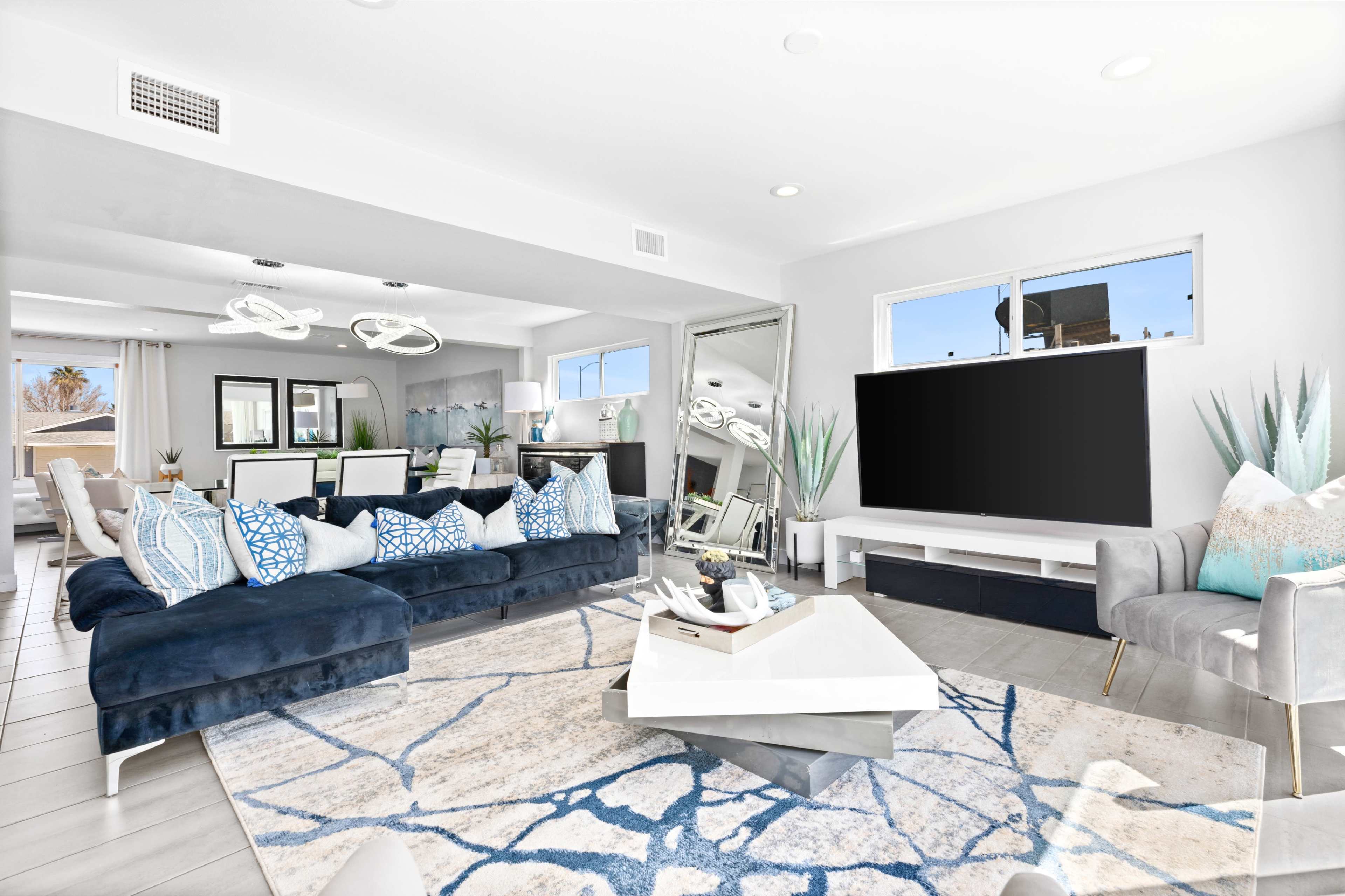The image shows a modern living room featuring a blue sectional sofa with patterned cushions, a large TV on a white media console, and a geometric rug on the tiled floor.