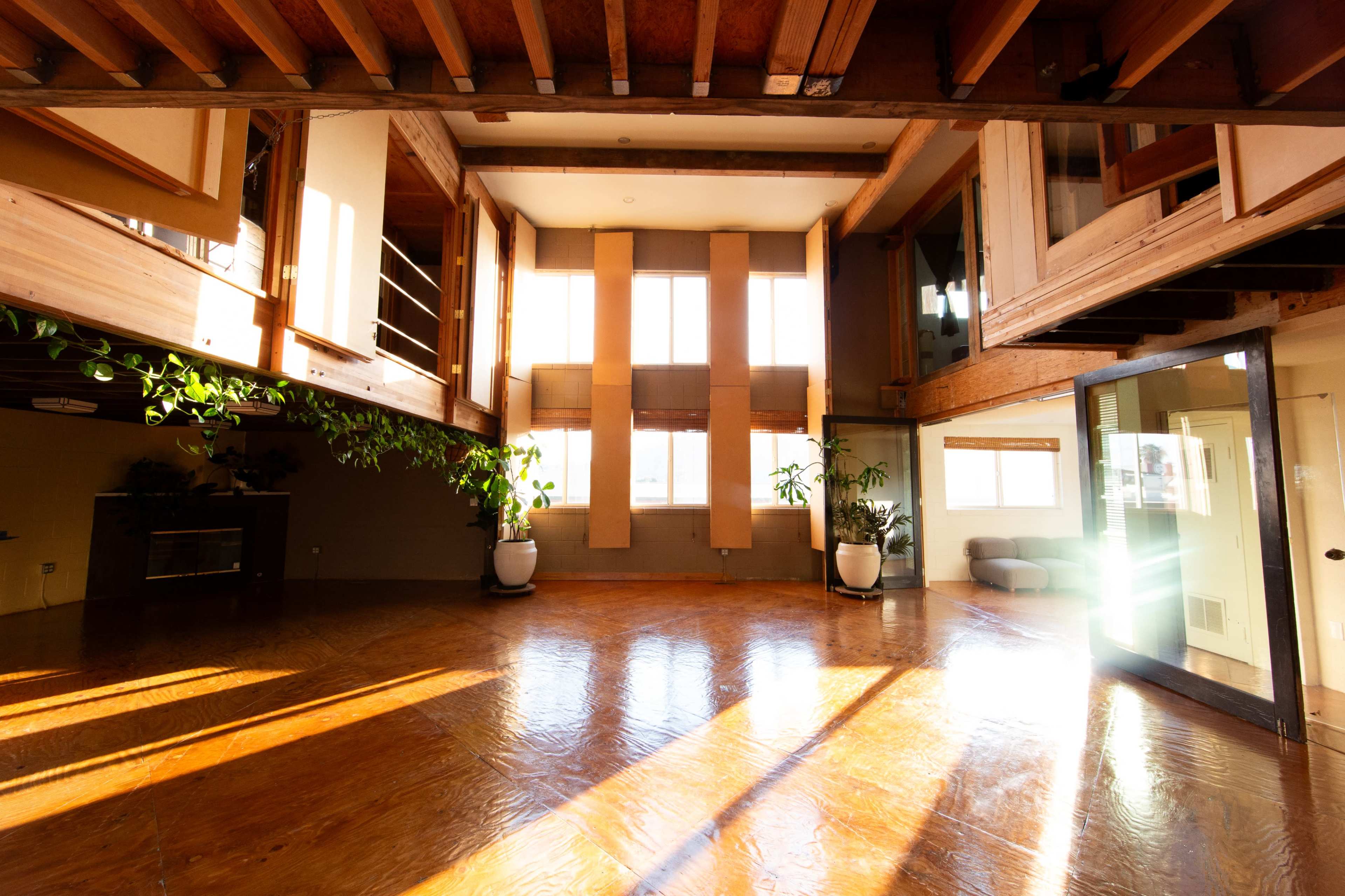 The interior of a spacious, modern living area features wooden beams, large windows, and potted plants, with sunlight illuminating the polished floor.