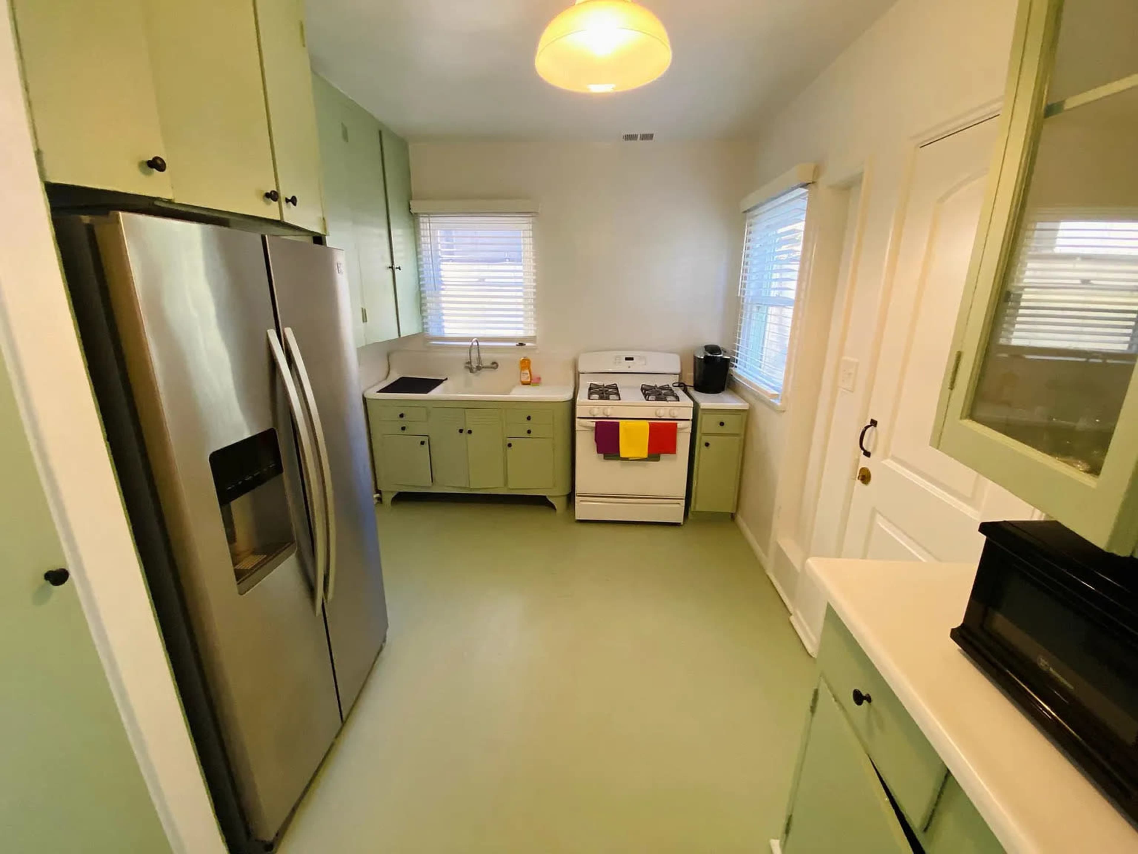 A small kitchen features light green cabinets, a stainless steel refrigerator, a white stove, and a sink near a window.