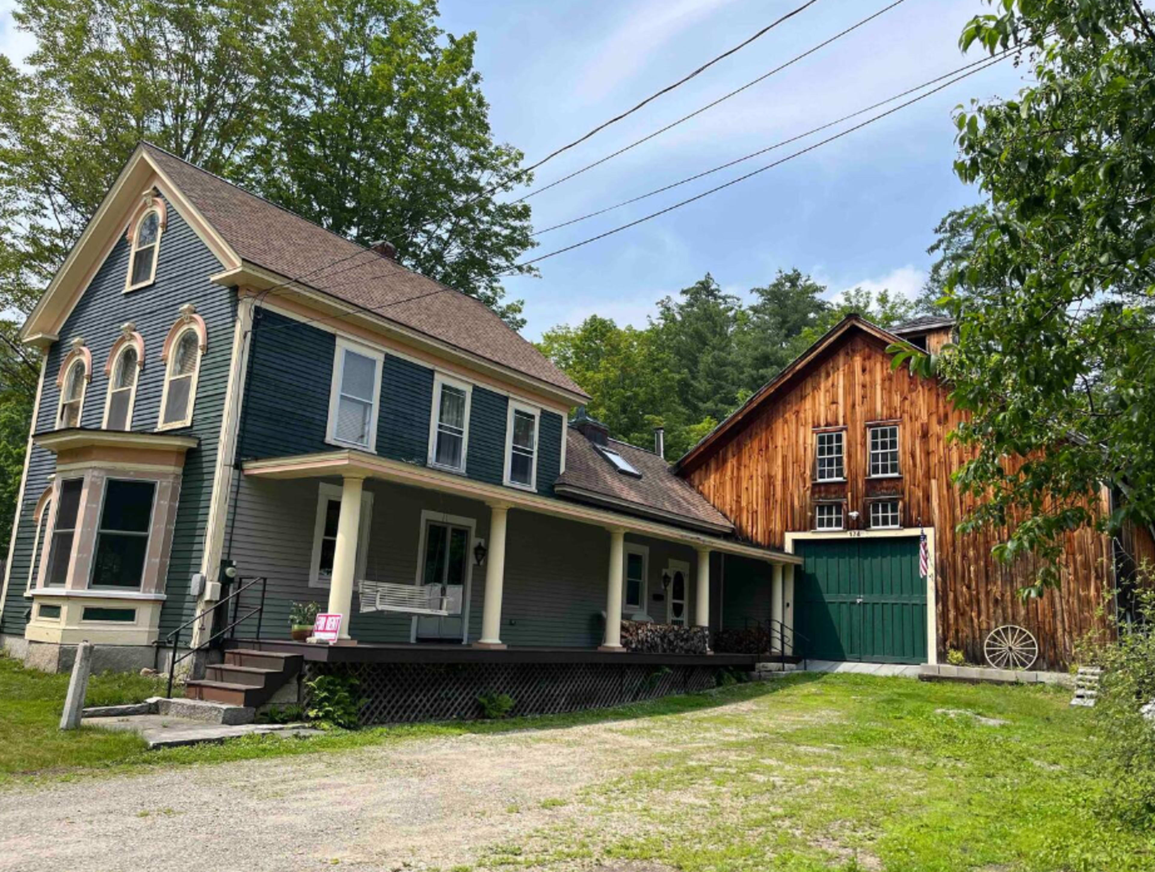 A two-story gray house with a covered porch beside a large wooden barn surrounded by trees.