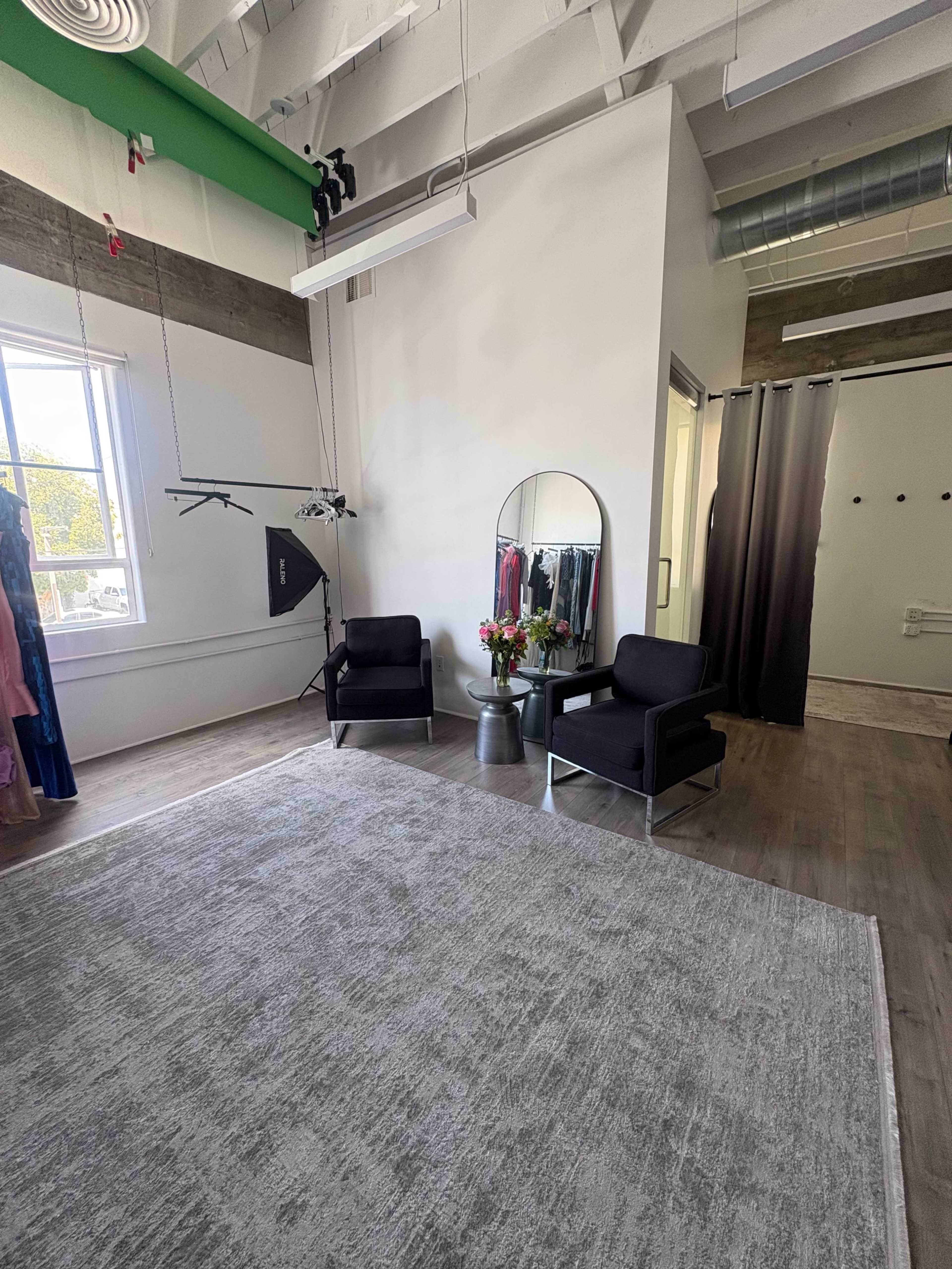 The image shows a spacious room with two black chairs, a large mirror, a floral arrangement, and colorful garments hanging on a rack.