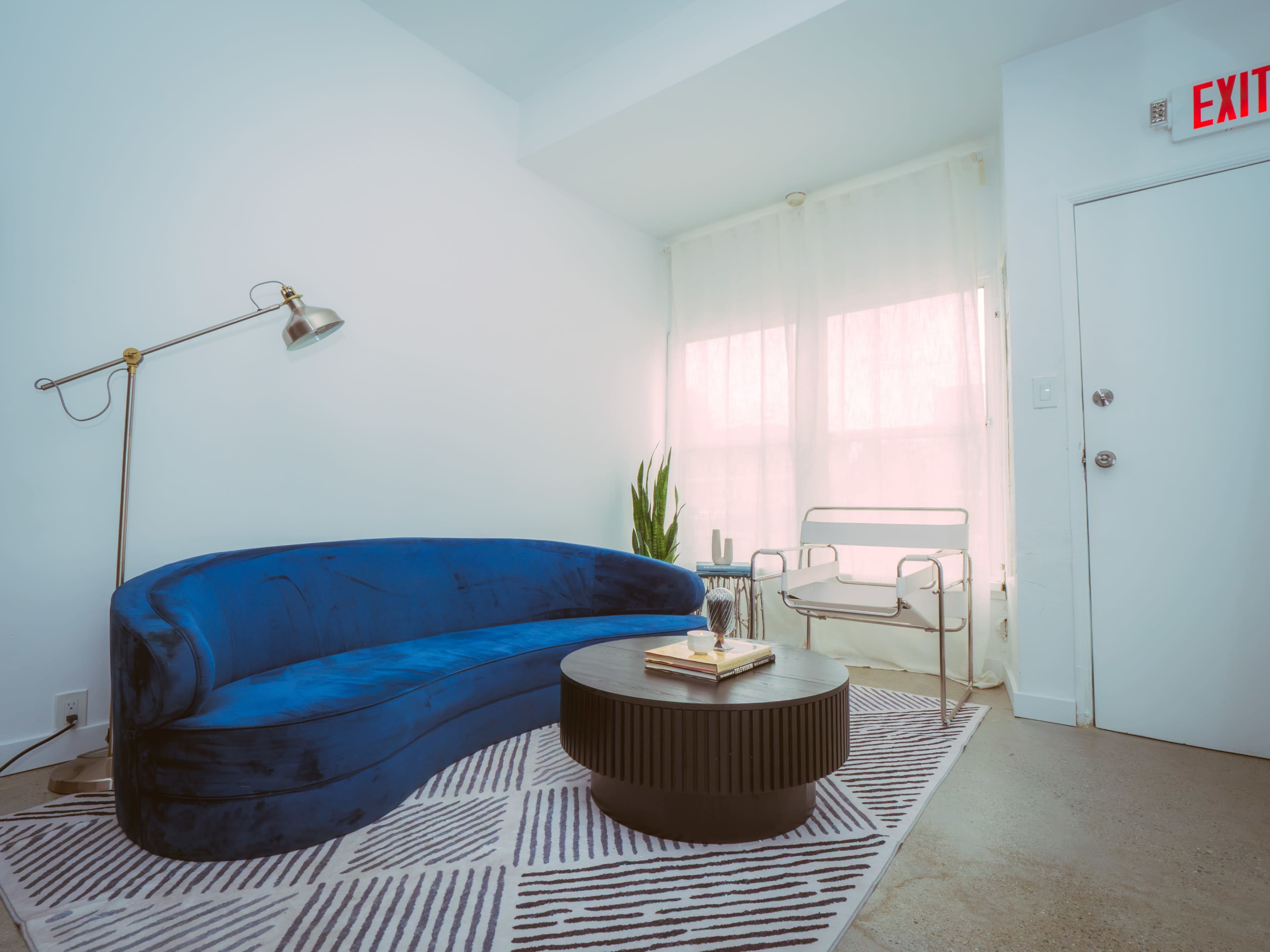 A modern living room features a blue velvet curved sofa, a round black coffee table, a clear chair, and a window with sheer curtains.