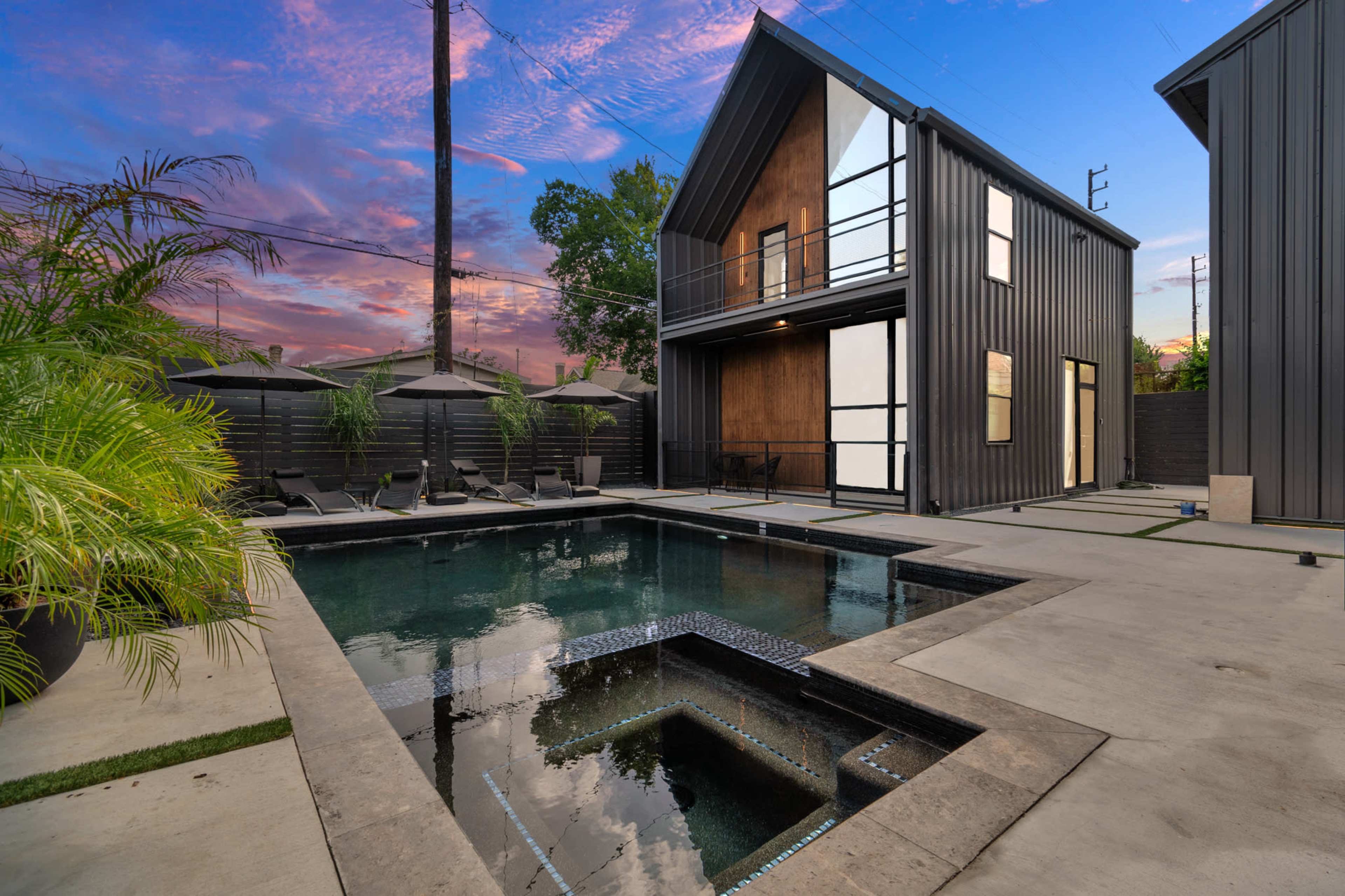 A modern, two-story house with sleek black exterior walls surrounding a rectangular swimming pool and a spa area.