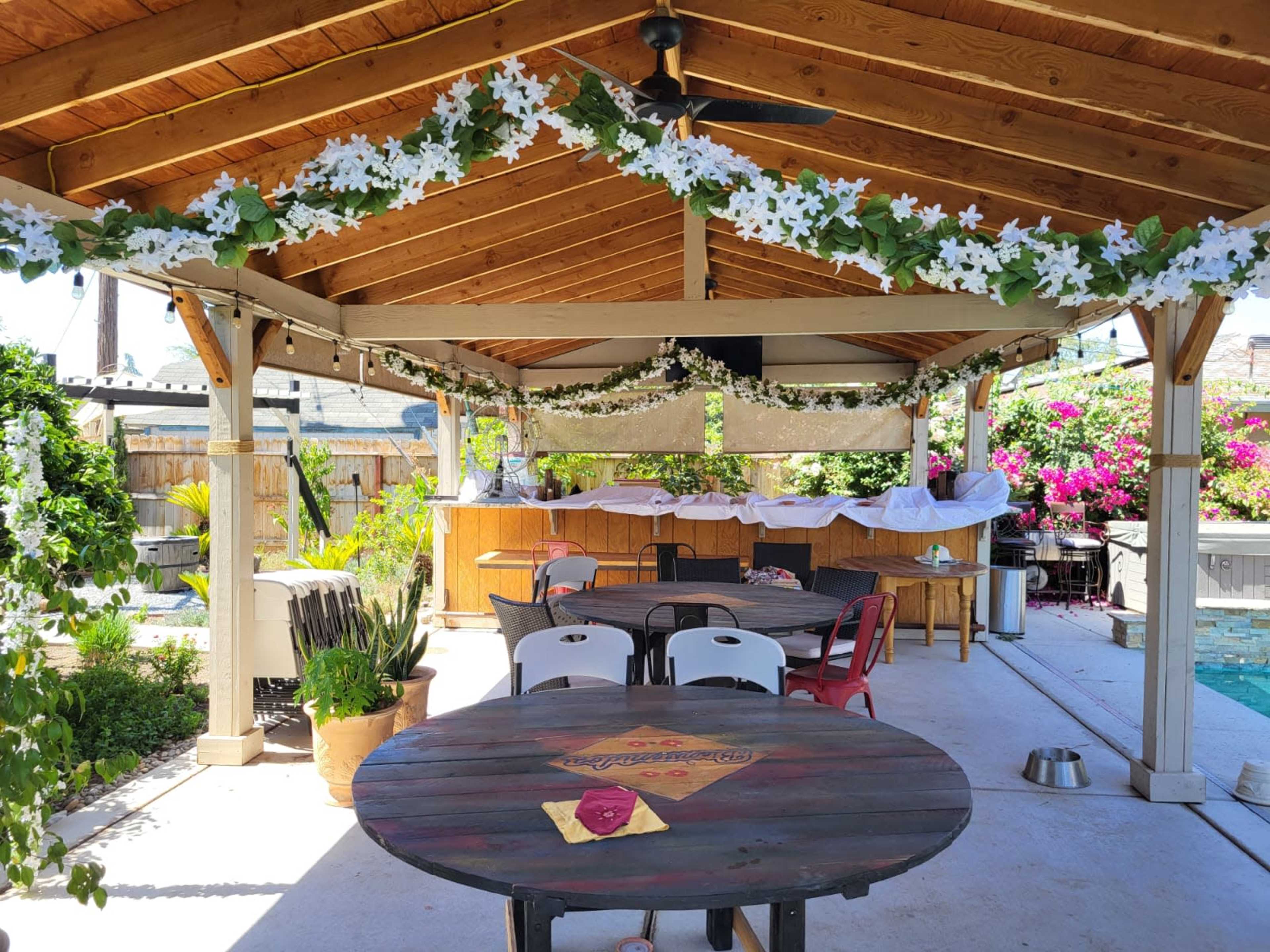 A covered outdoor space features a wooden ceiling with floral decorations, tables, and a pool in the background.