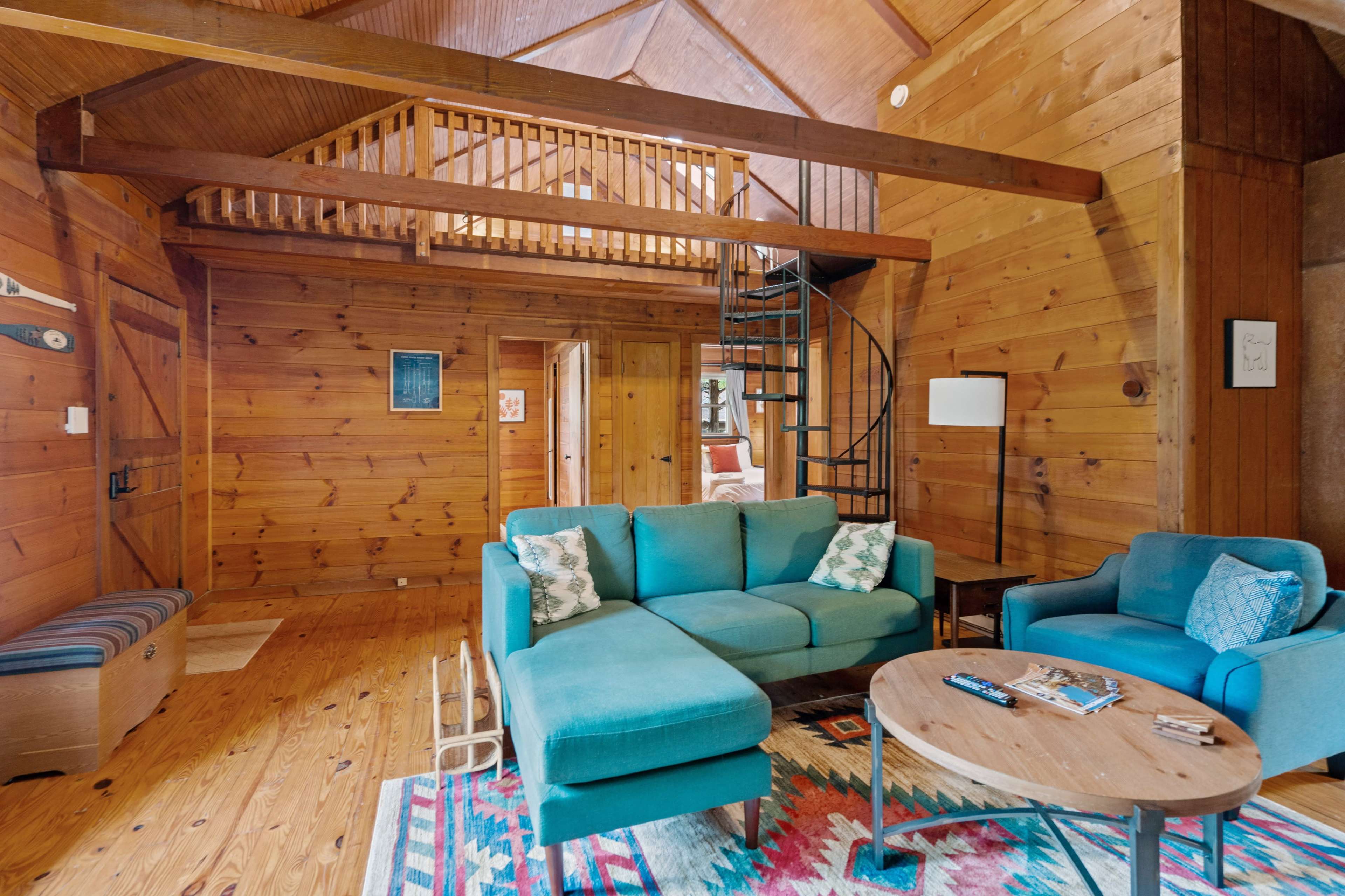 A wooden cabin interior with a spiral staircase, a teal sectional sofa, and a round coffee table on a colorful rug.