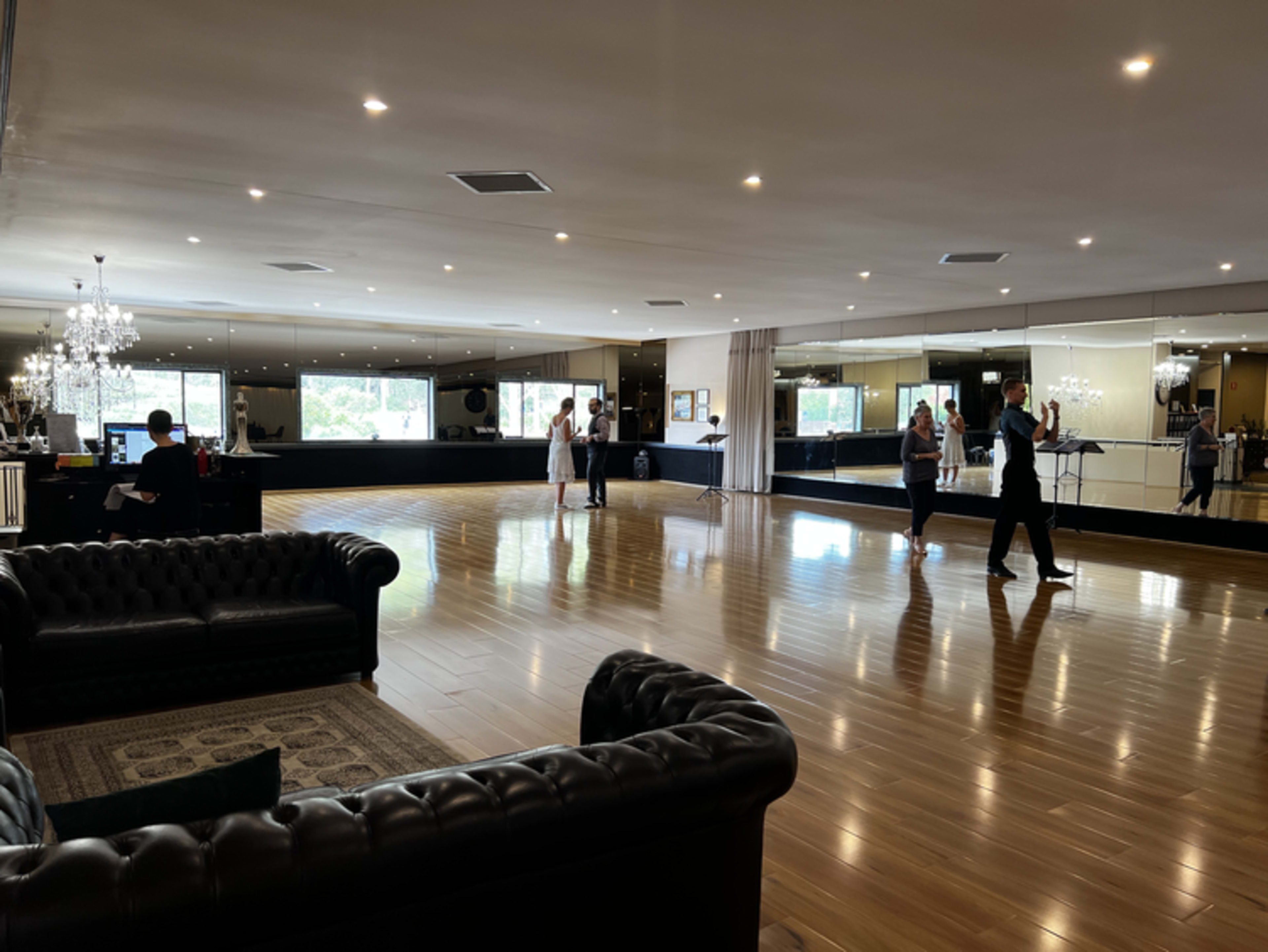 A spacious dance studio features polished wooden floors, large mirrors, and couples practicing dancing while others observe.