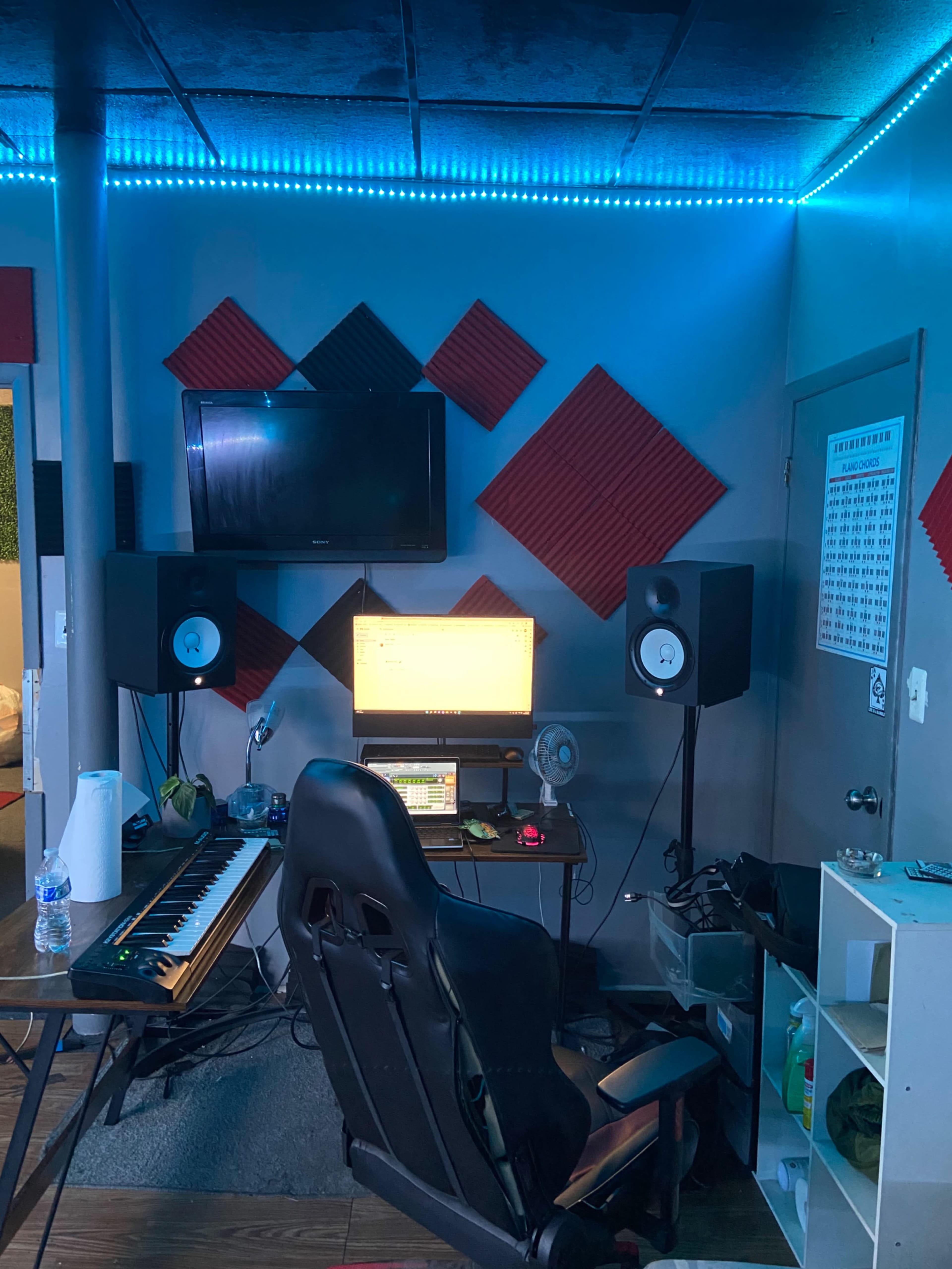 A music production studio with a computer setup, audio speakers, a MIDI keyboard, and acoustic foam panels on the walls.