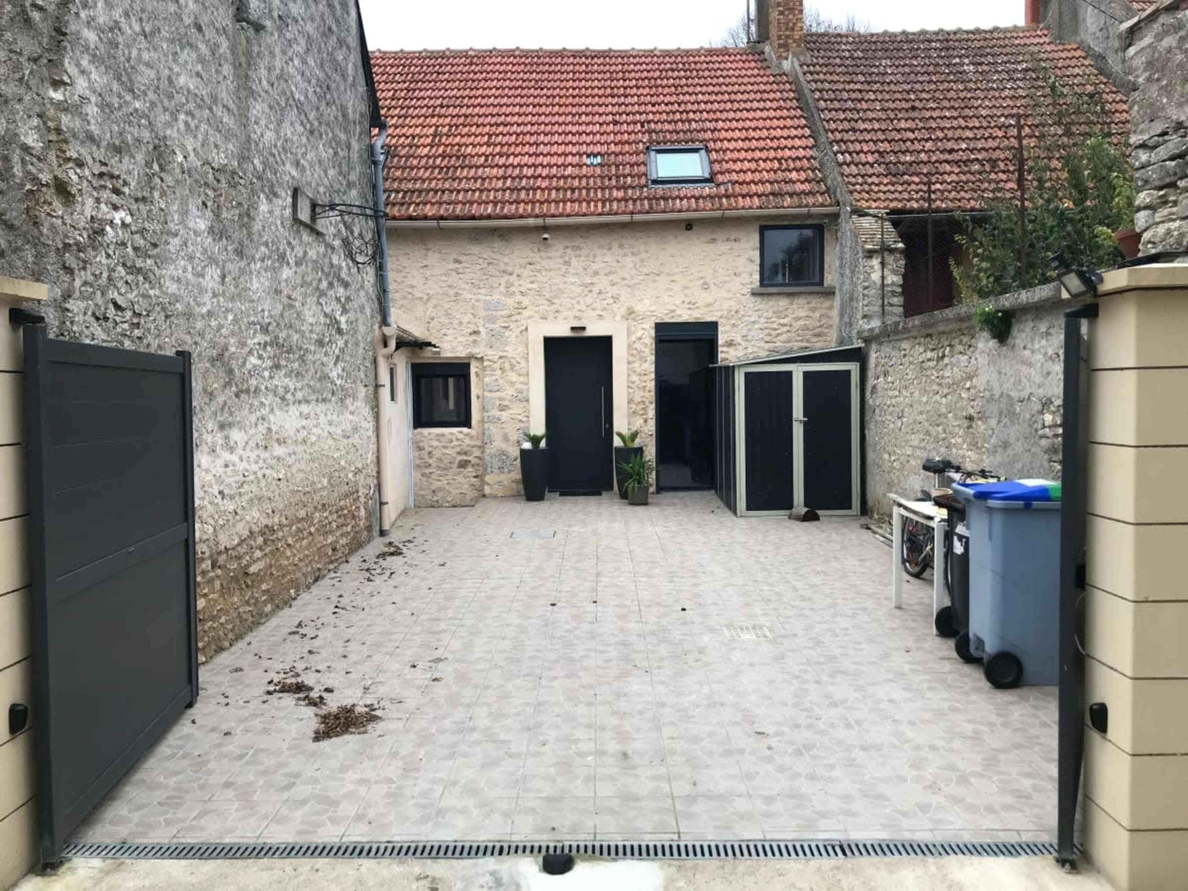 The image shows a paved outdoor space at the back of a house with a stone facade, featuring a small garden and several utility bins.