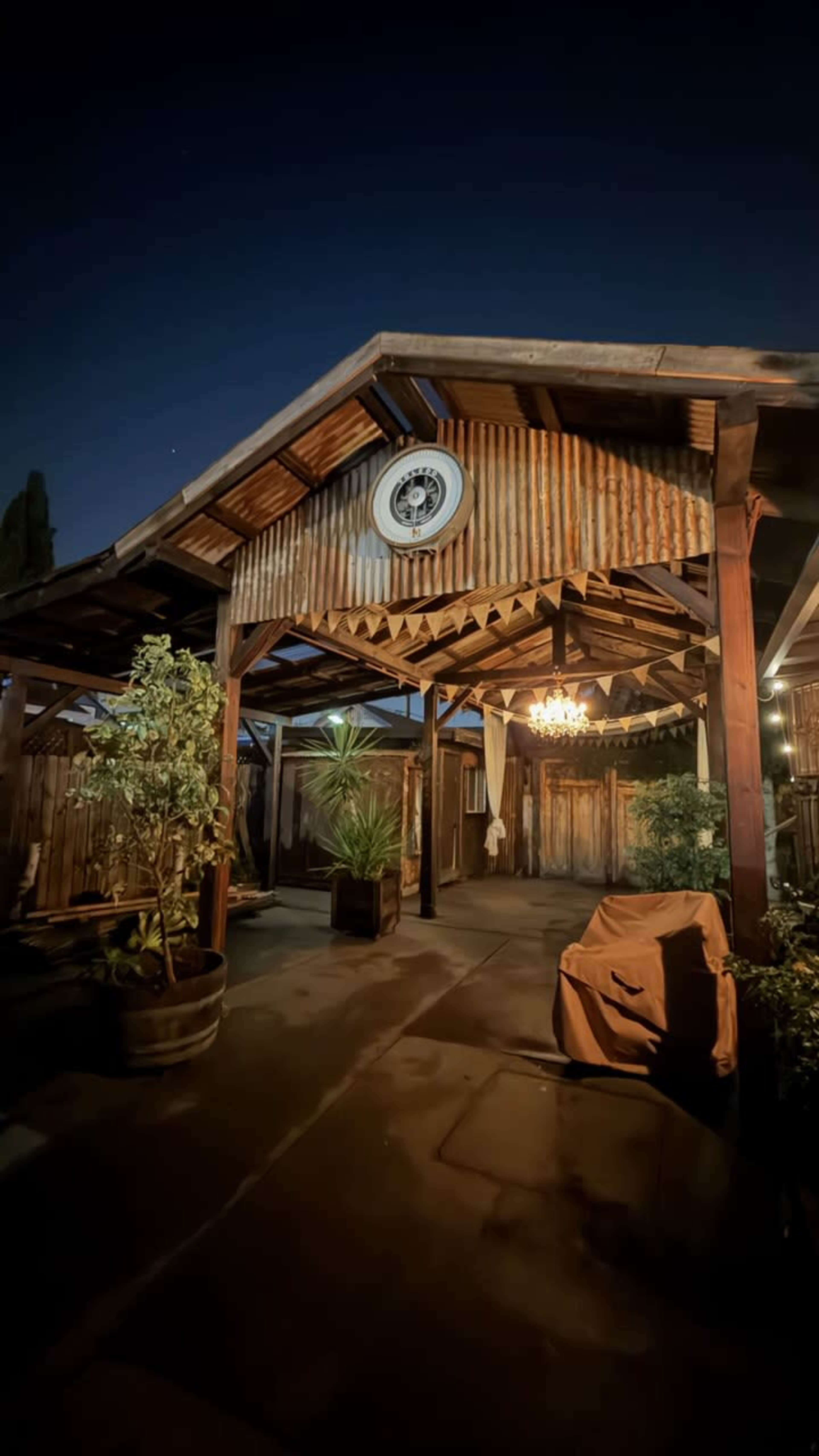The image shows a rustic outdoor patio with a corrugated metal roof, wooden beams, potted plants, and a chandelier hanging from the ceiling.