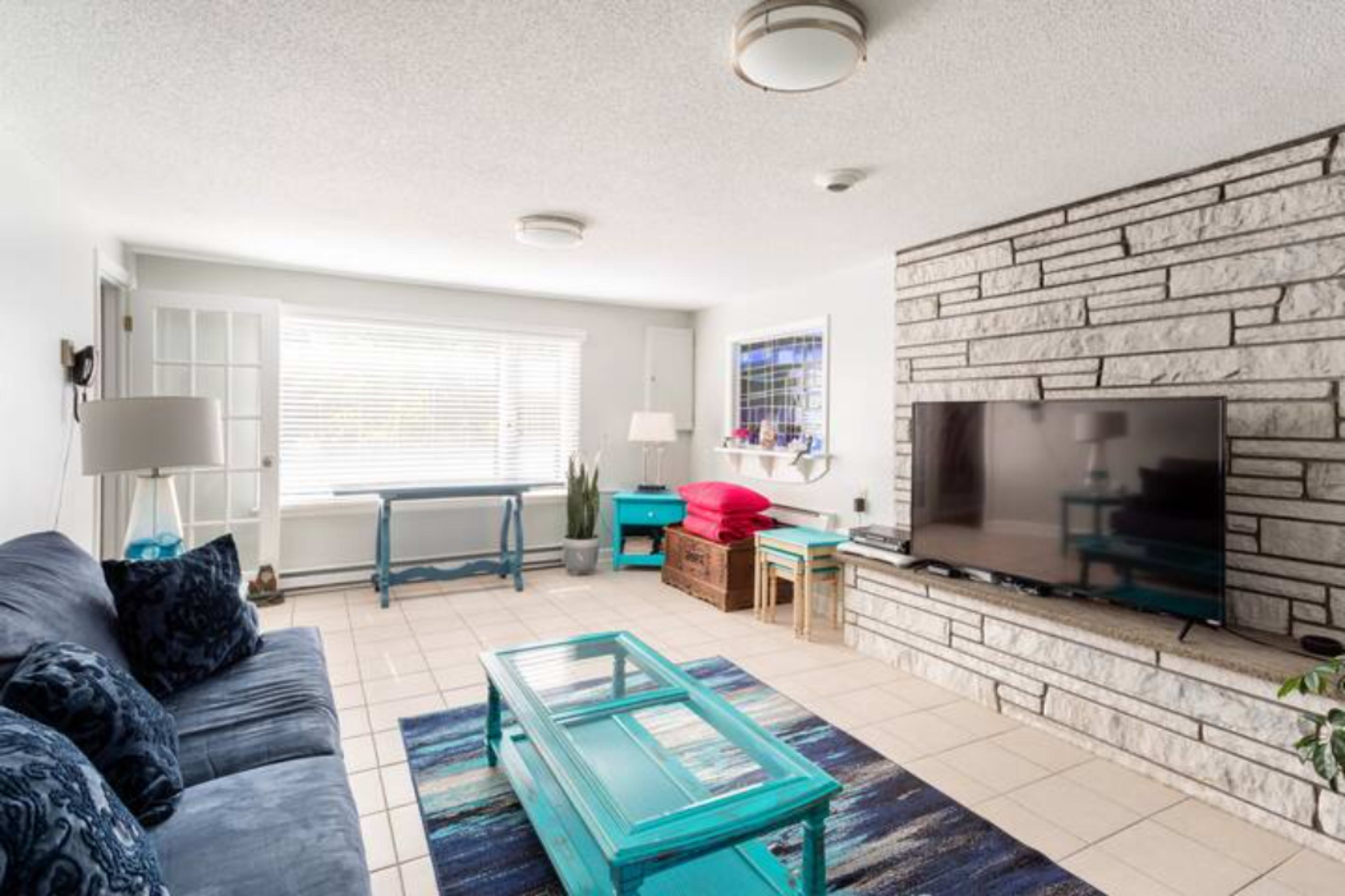 The living room features a blue sofa, a glass coffee table, a stone accent wall, and a large television accompanied by colorful furniture and decor.