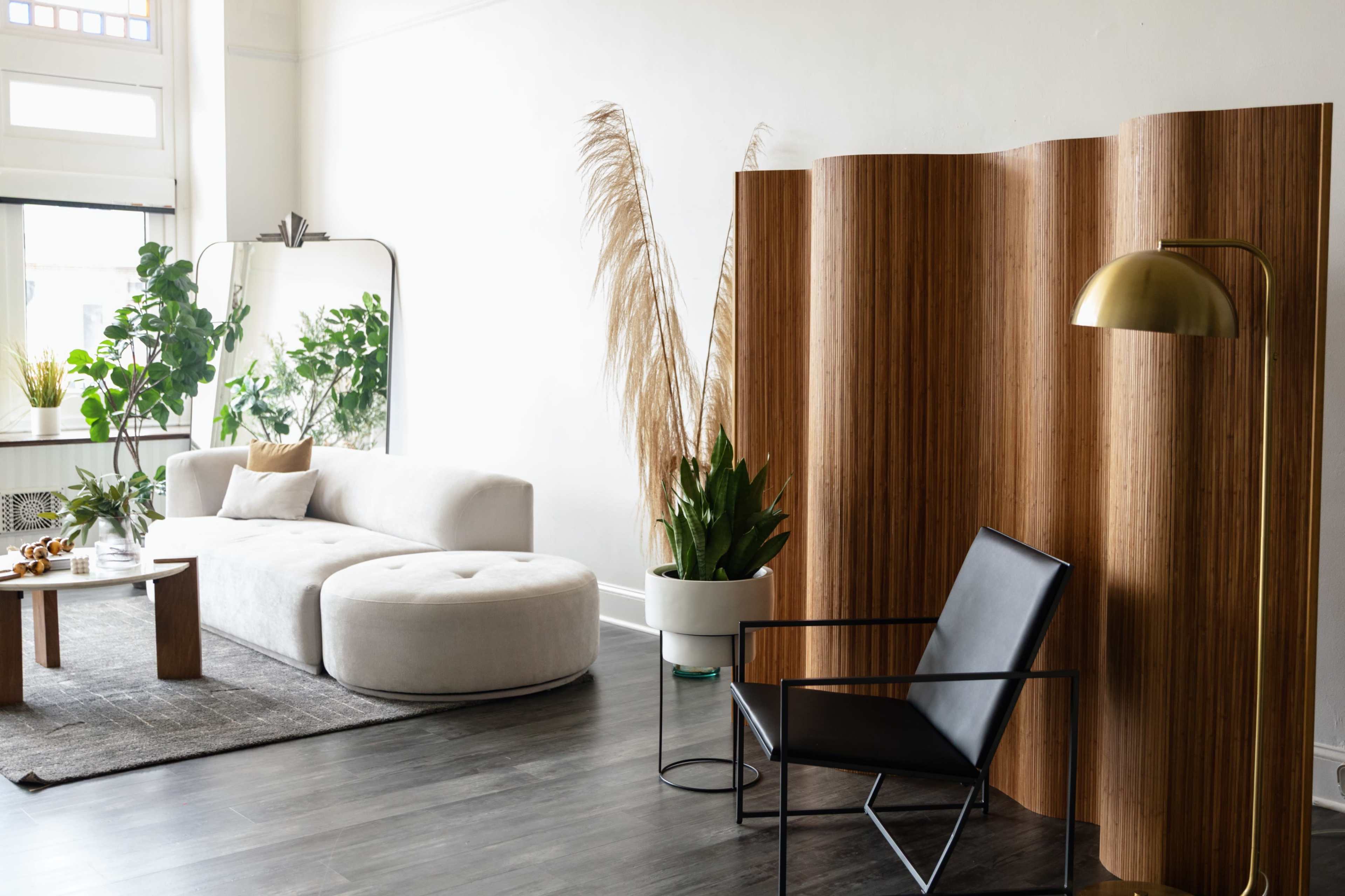 The image shows a modern living room featuring a large round sofa, a black chair, a wooden coffee table, and potted plants, all set against a light-colored wall.