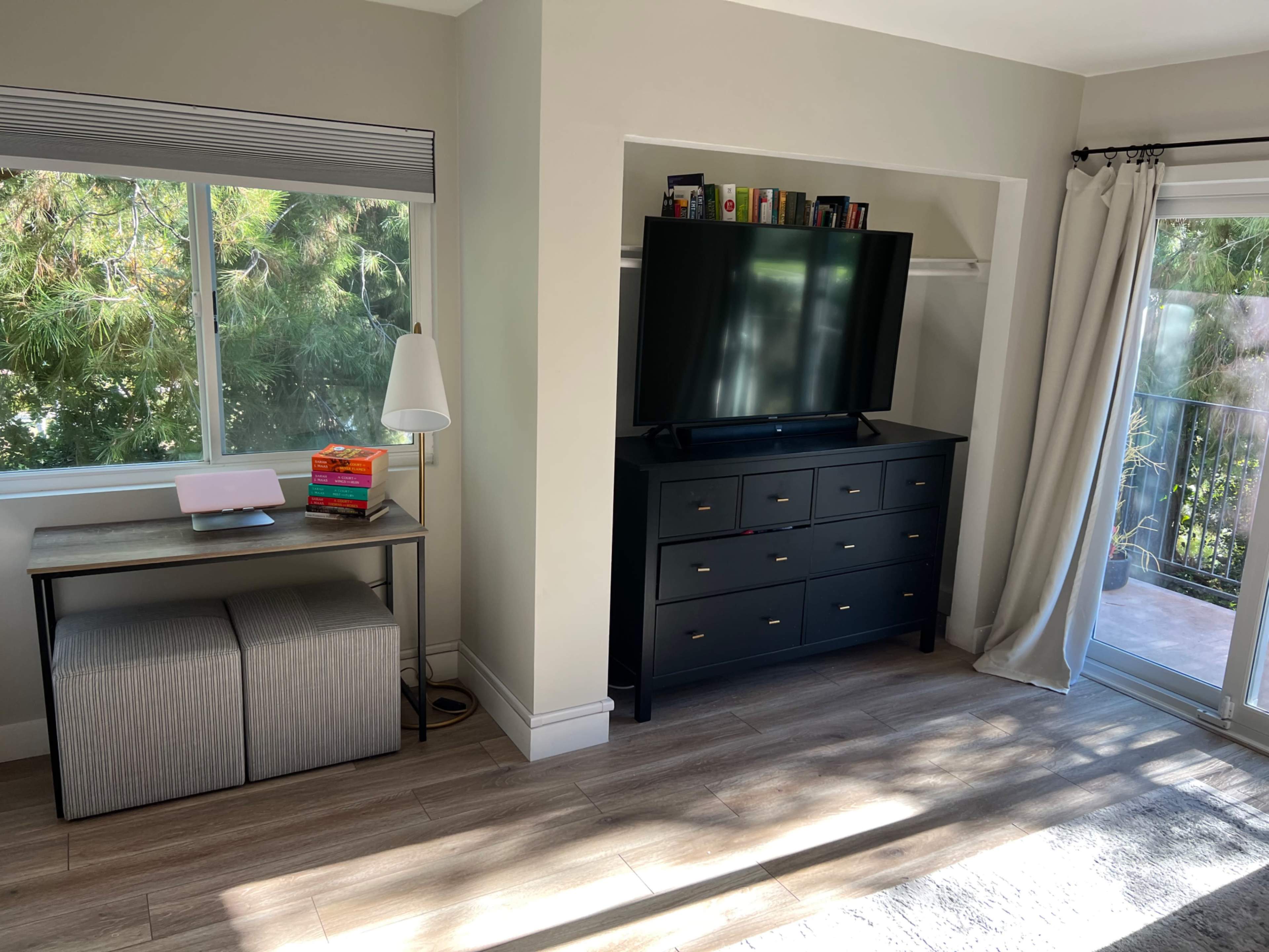 A living space features a dark dresser with a television on top, a small desk with a laptop, and large windows overlooking greenery.