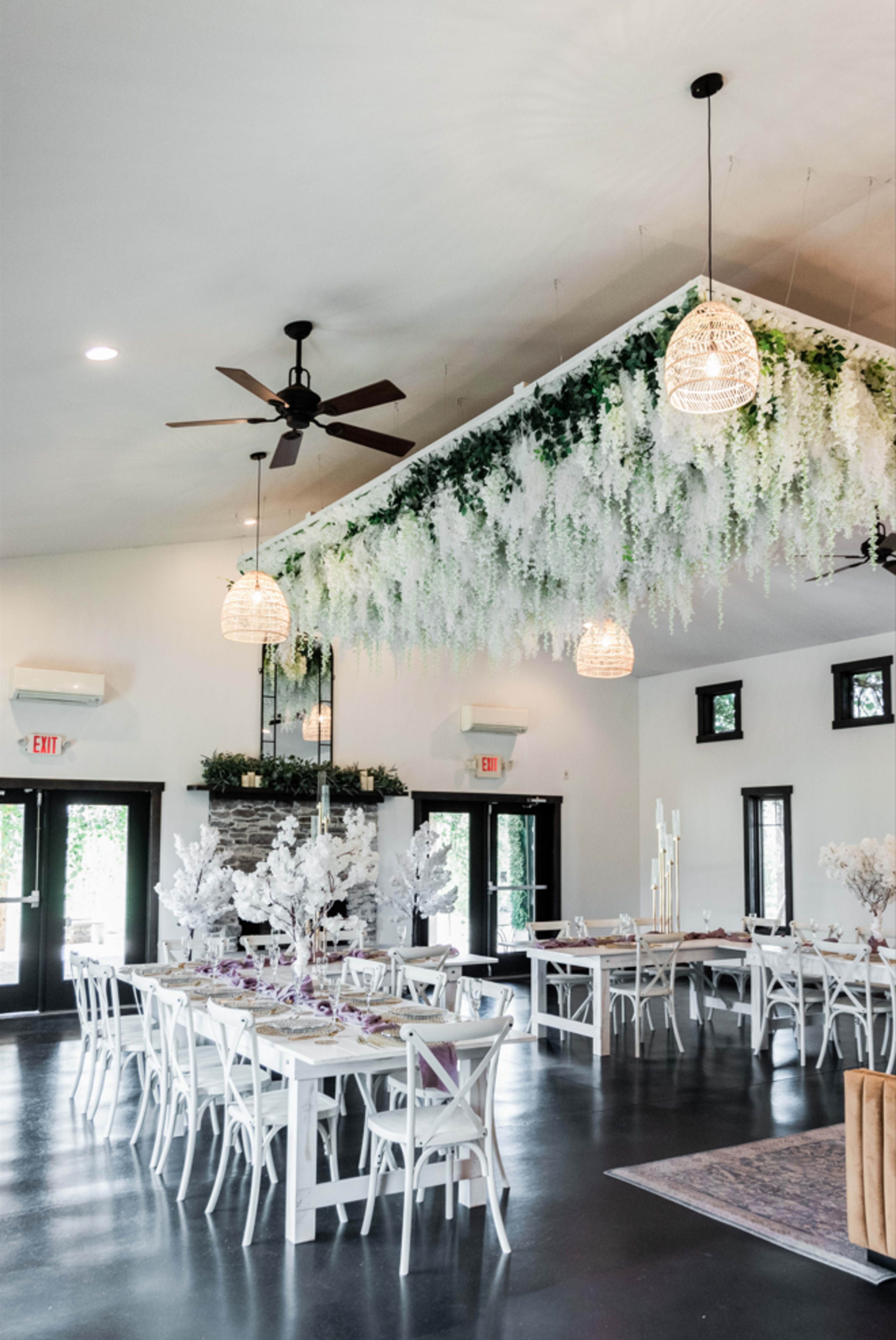 The interior of a spacious event venue features tables with white chairs, adorned with floral decorations, and a ceiling installation of greenery and hanging pendant lights.