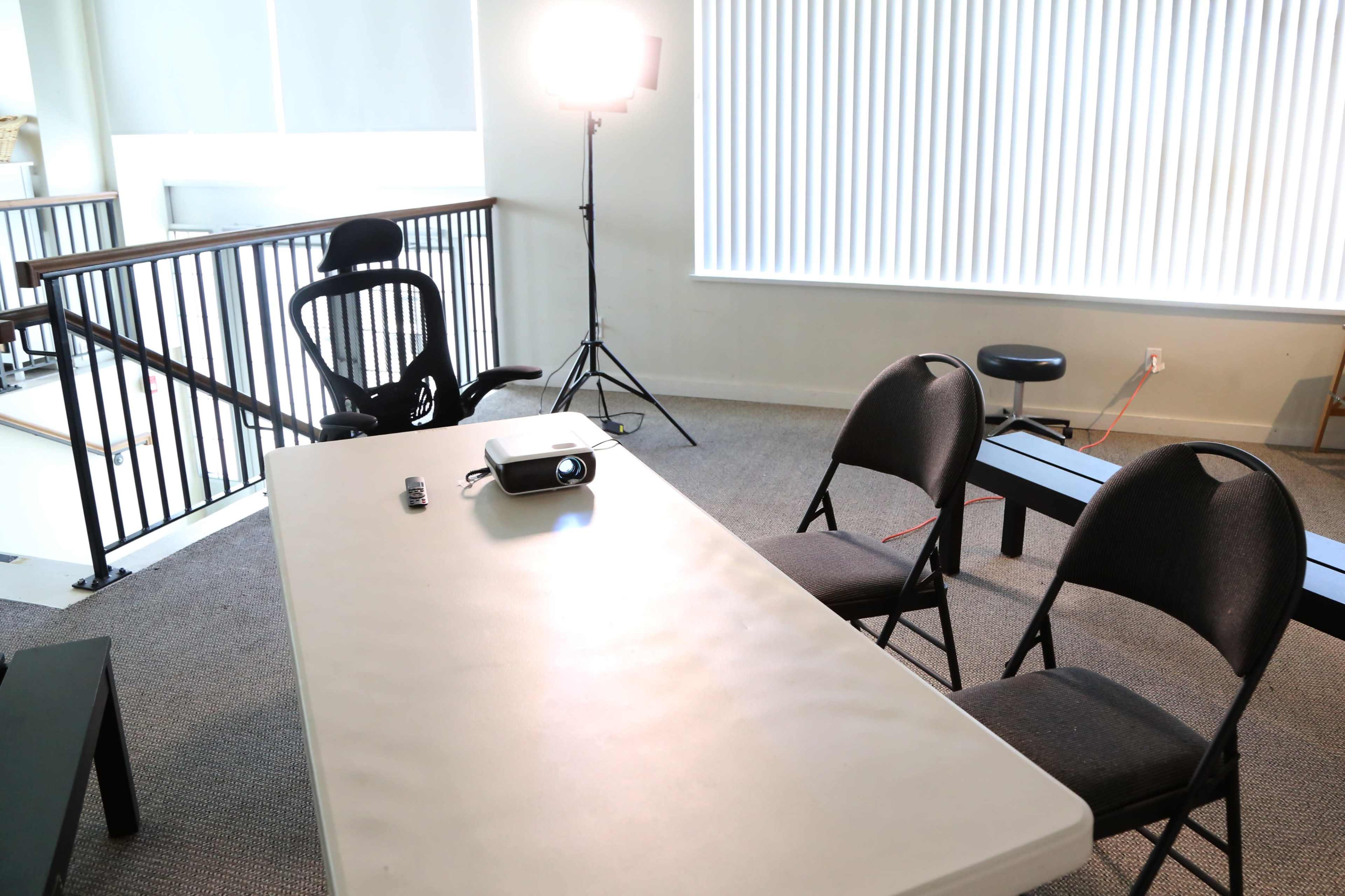 The scene shows a well-lit room featuring a table with a projector, an office chair, and two folding chairs arranged around it.