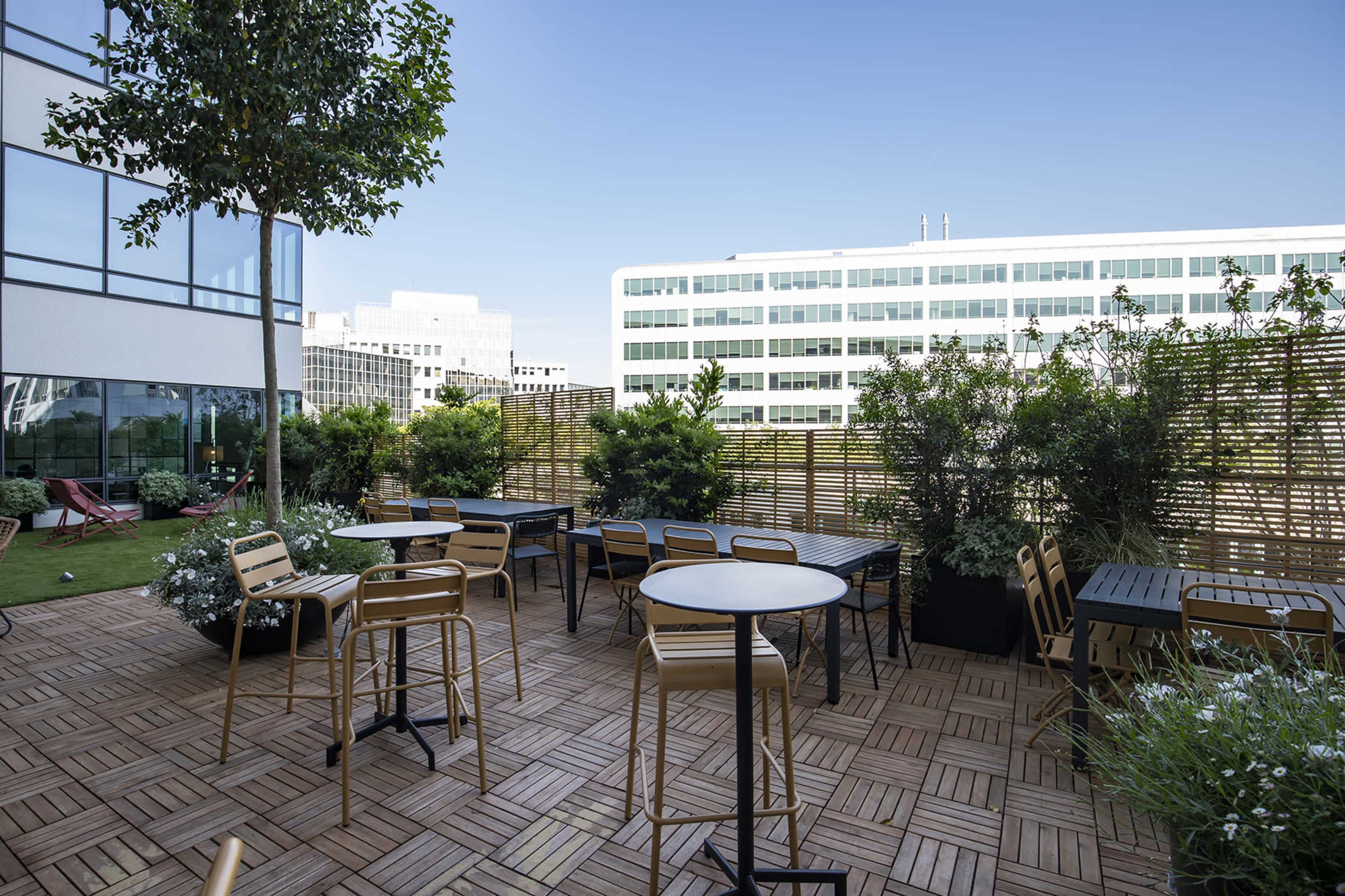 An outdoor terrace area with wooden floors, tables, and chairs surrounded by greenery and tall buildings in the background.