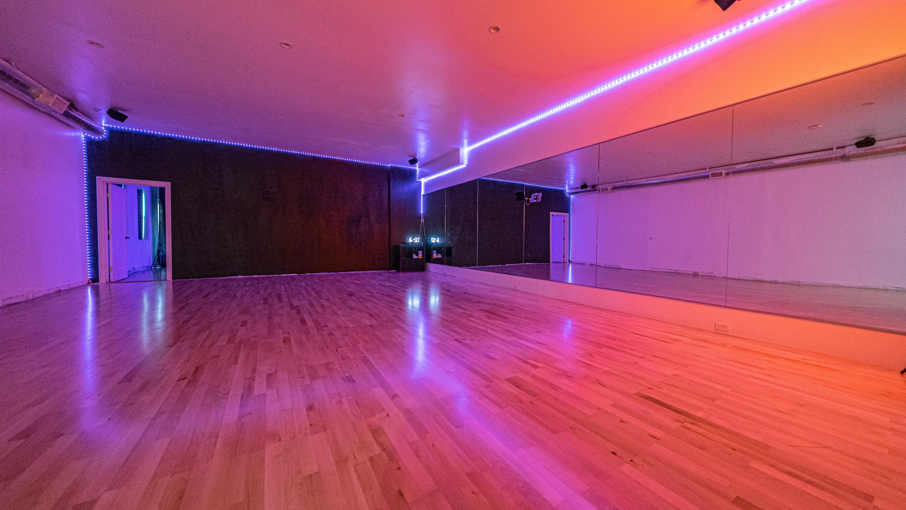 A spacious dance studio features polished wooden floors, mirrored walls, and colorful LED lighting along the perimeter.