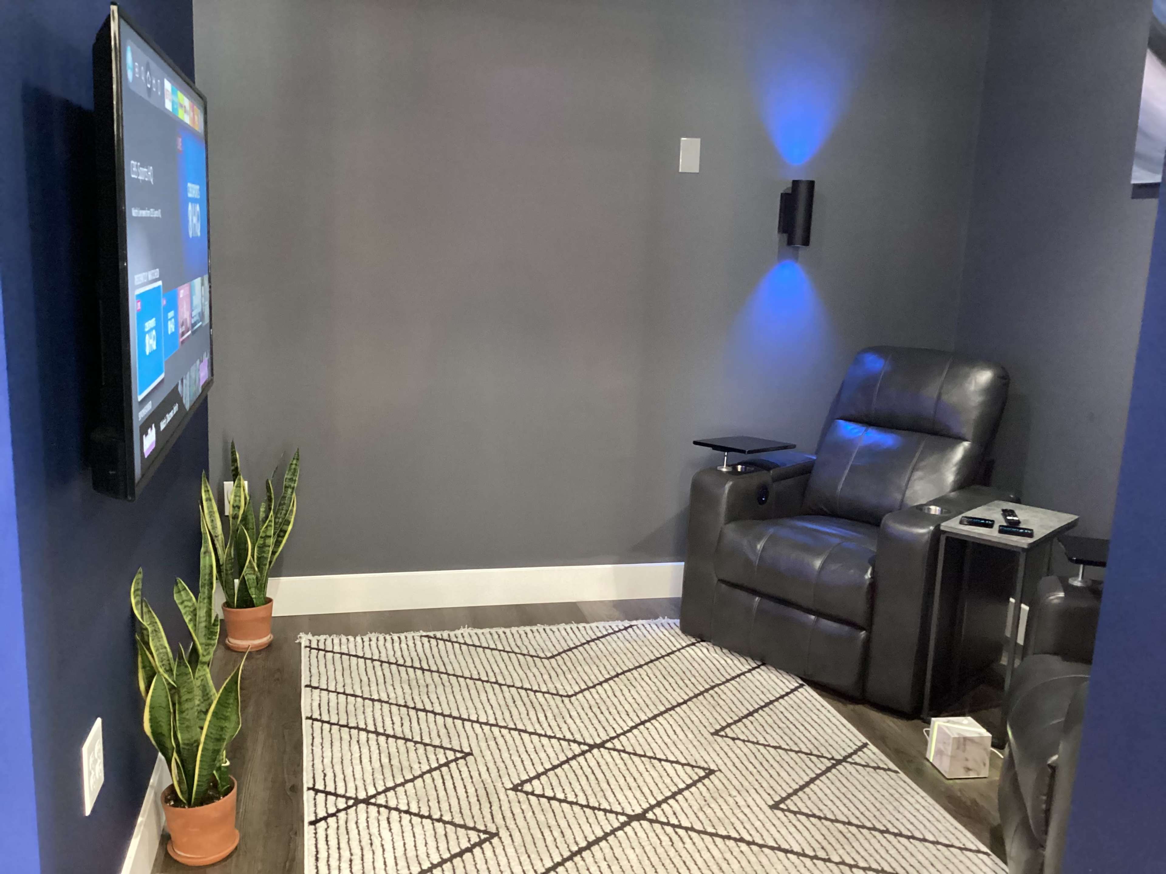 A cozy room with a recliner chair facing a wall-mounted television, flanked by green plants on either side and a patterned rug on the floor.