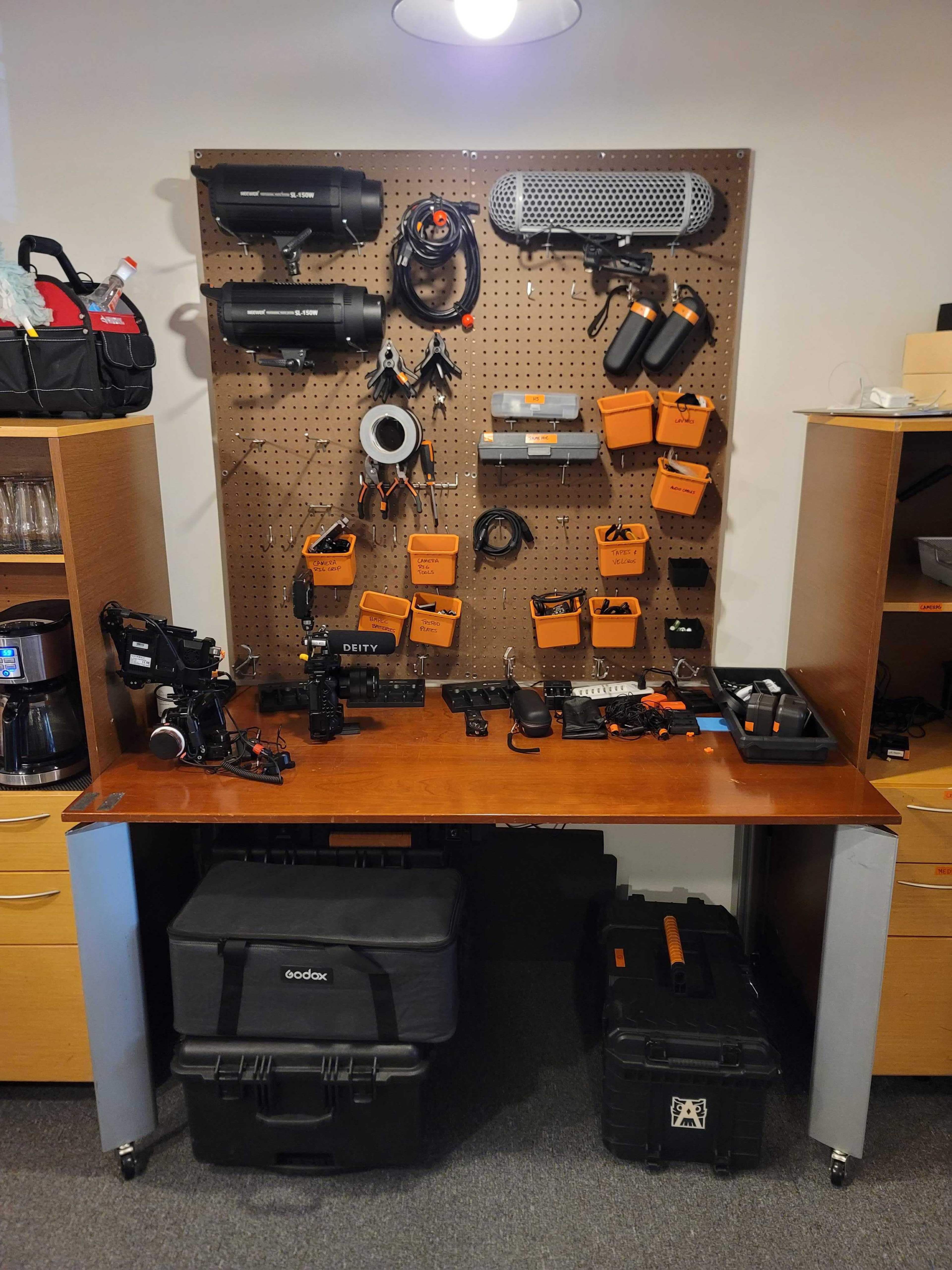 A wooden table with various photography and videography equipment organized on a pegboard above it, including lights, microphones, and camera accessories.