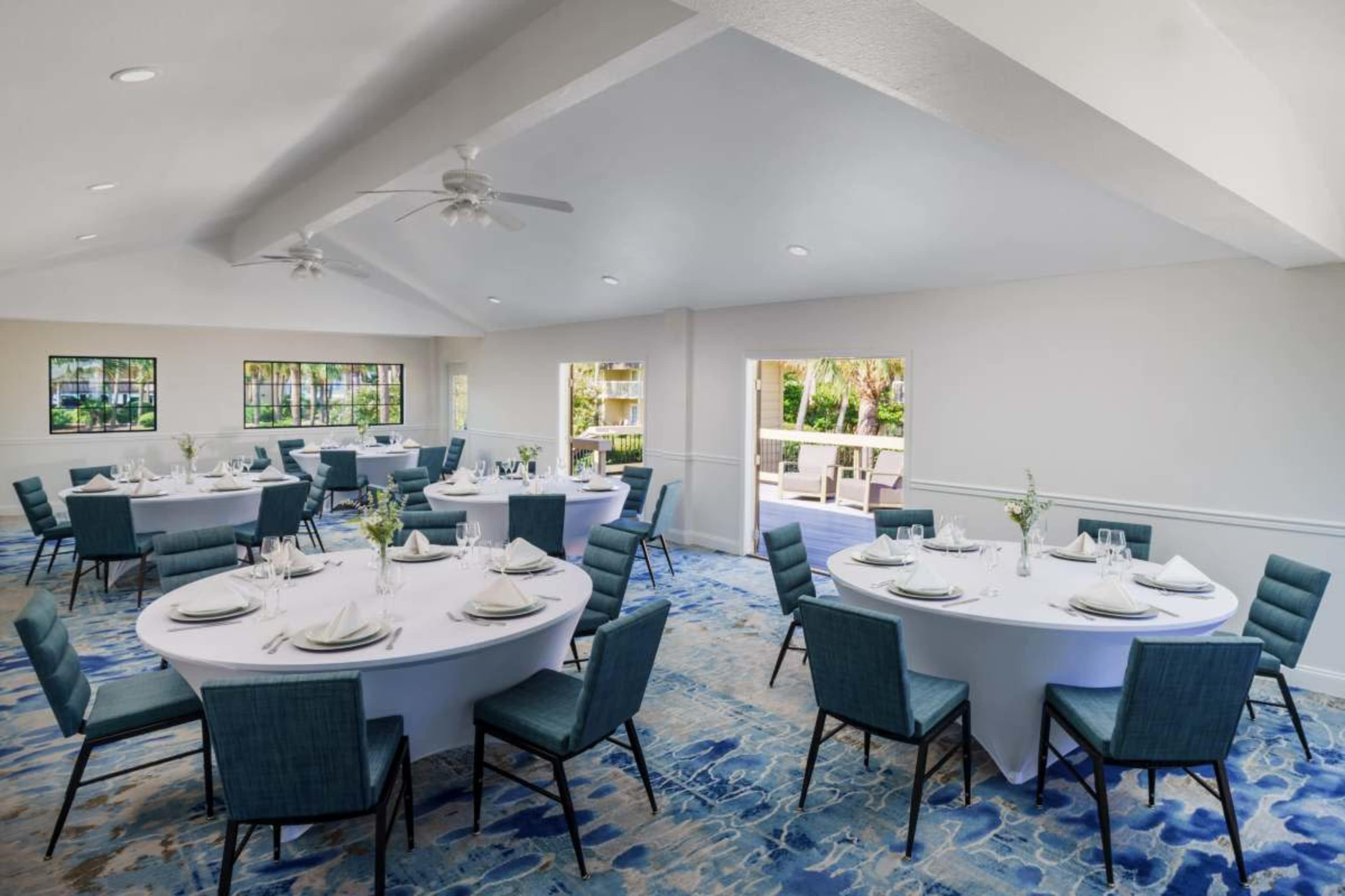 The image shows a bright, modern event space with two round dining tables set for a meal, surrounded by blue chairs and large windows that open to a view of an outdoor area.
