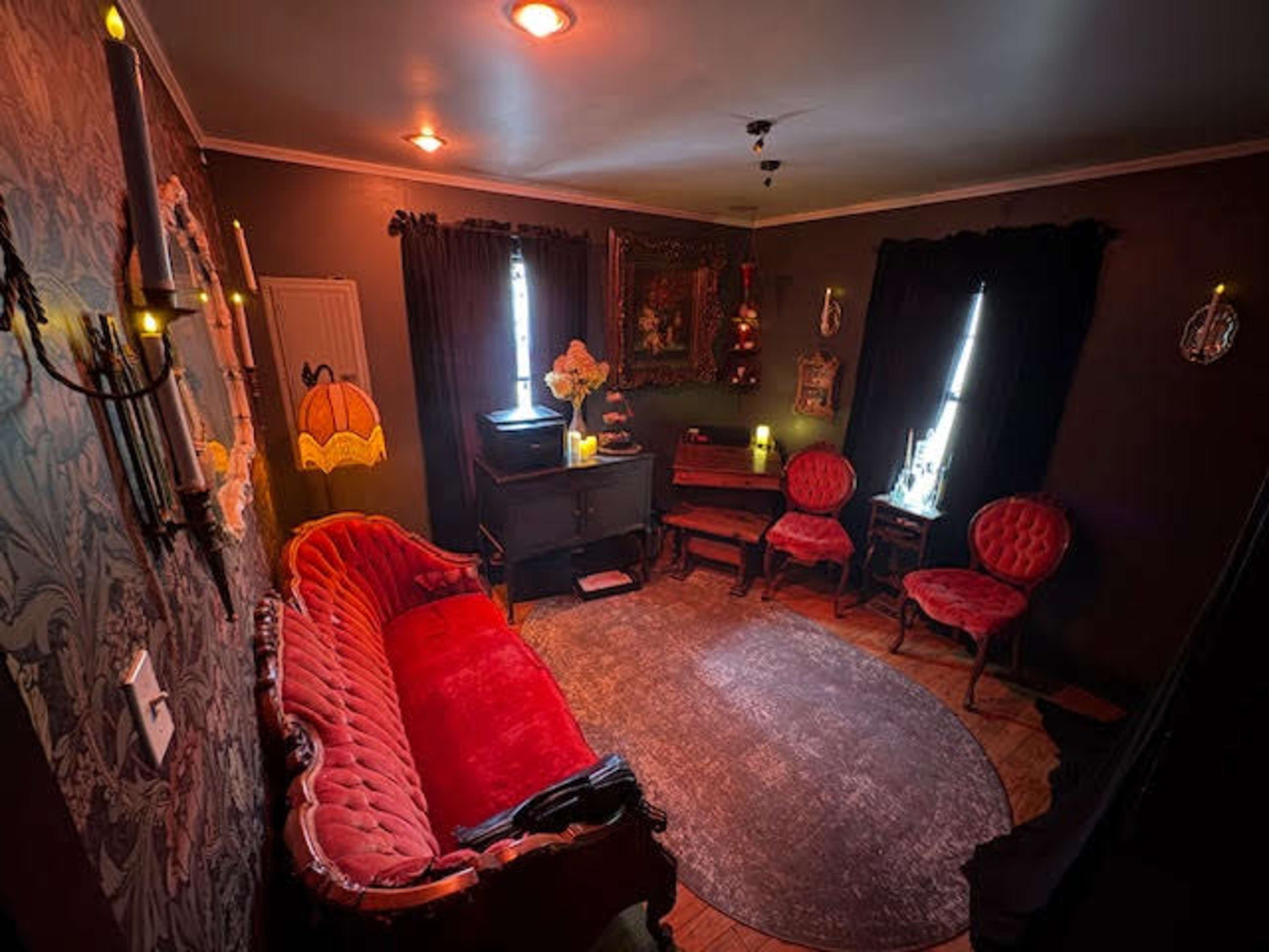 The room features a vintage red velvet sofa, two matching chairs, and a piano, all set against dark walls adorned with decorative elements.