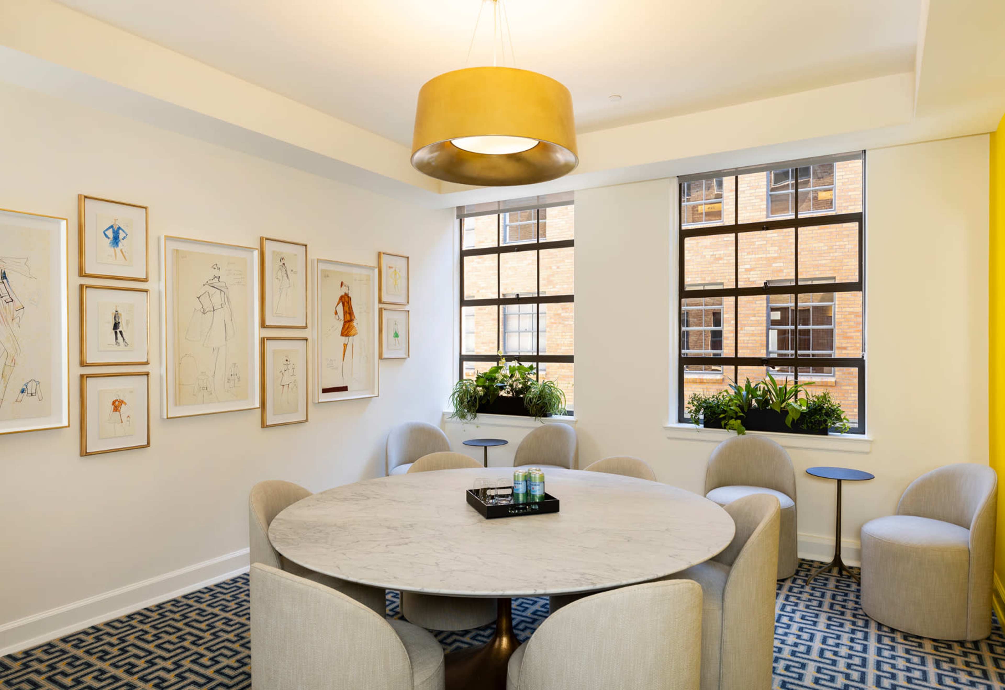 A circular marble table surrounded by upholstered chairs is situated in a well-lit room with large windows and framed artwork on the walls.