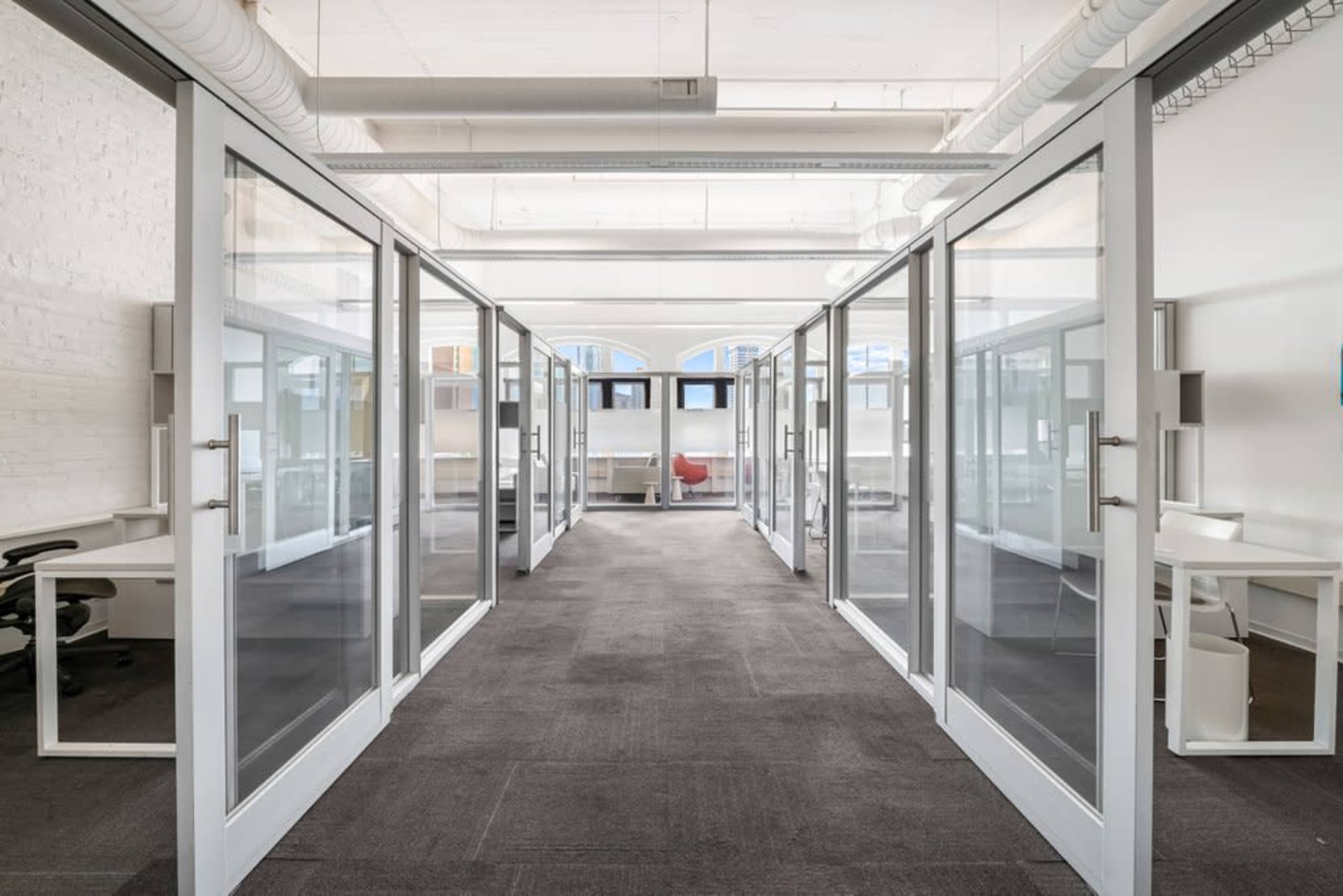 The image shows a modern office space featuring multiple glass-walled meeting rooms and workstations arranged along a central corridor.