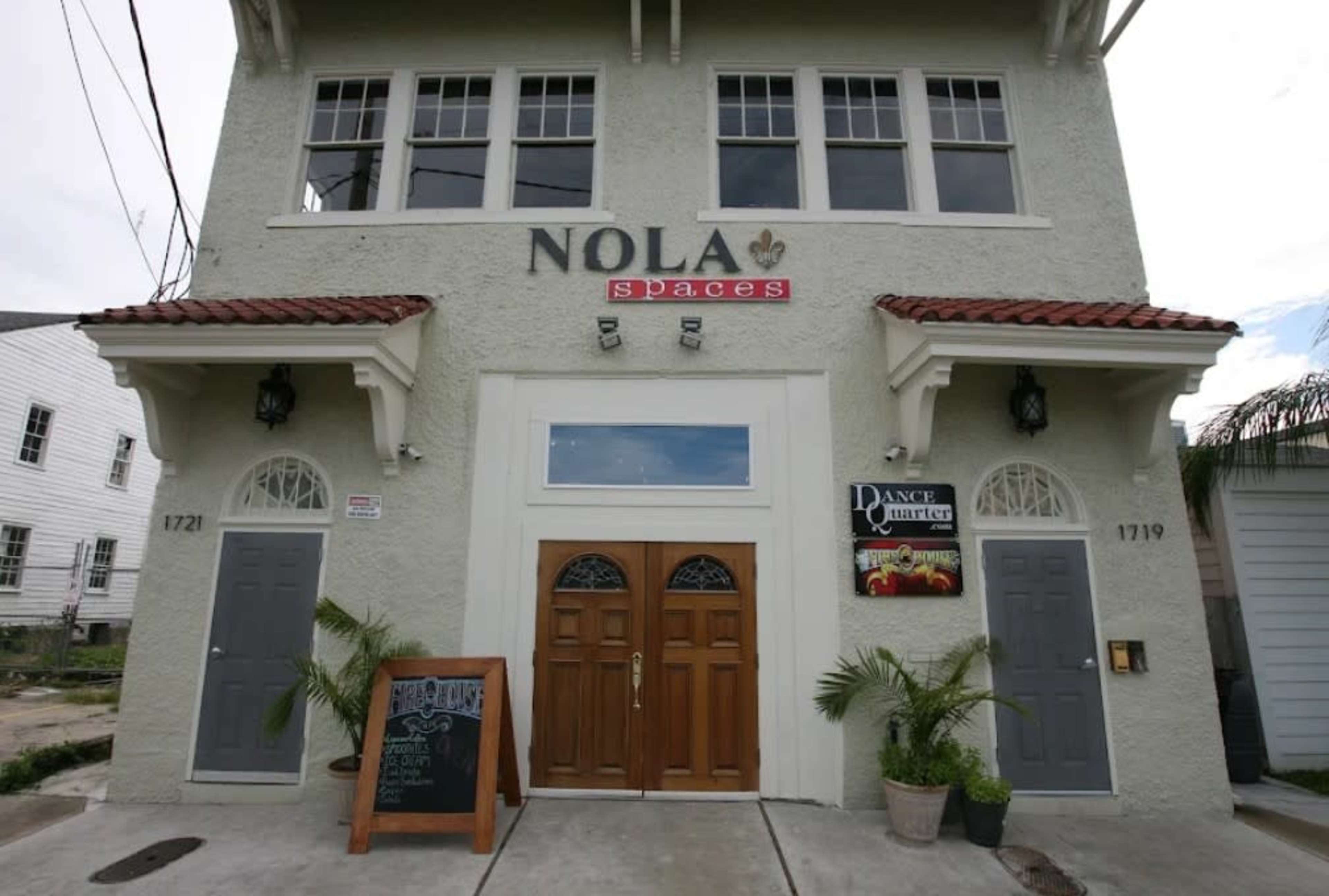 The image shows a two-story building with a sign reading "NOLA Spaces" above the entrance, featuring dual wooden doors flanked by large windows and two side doors.