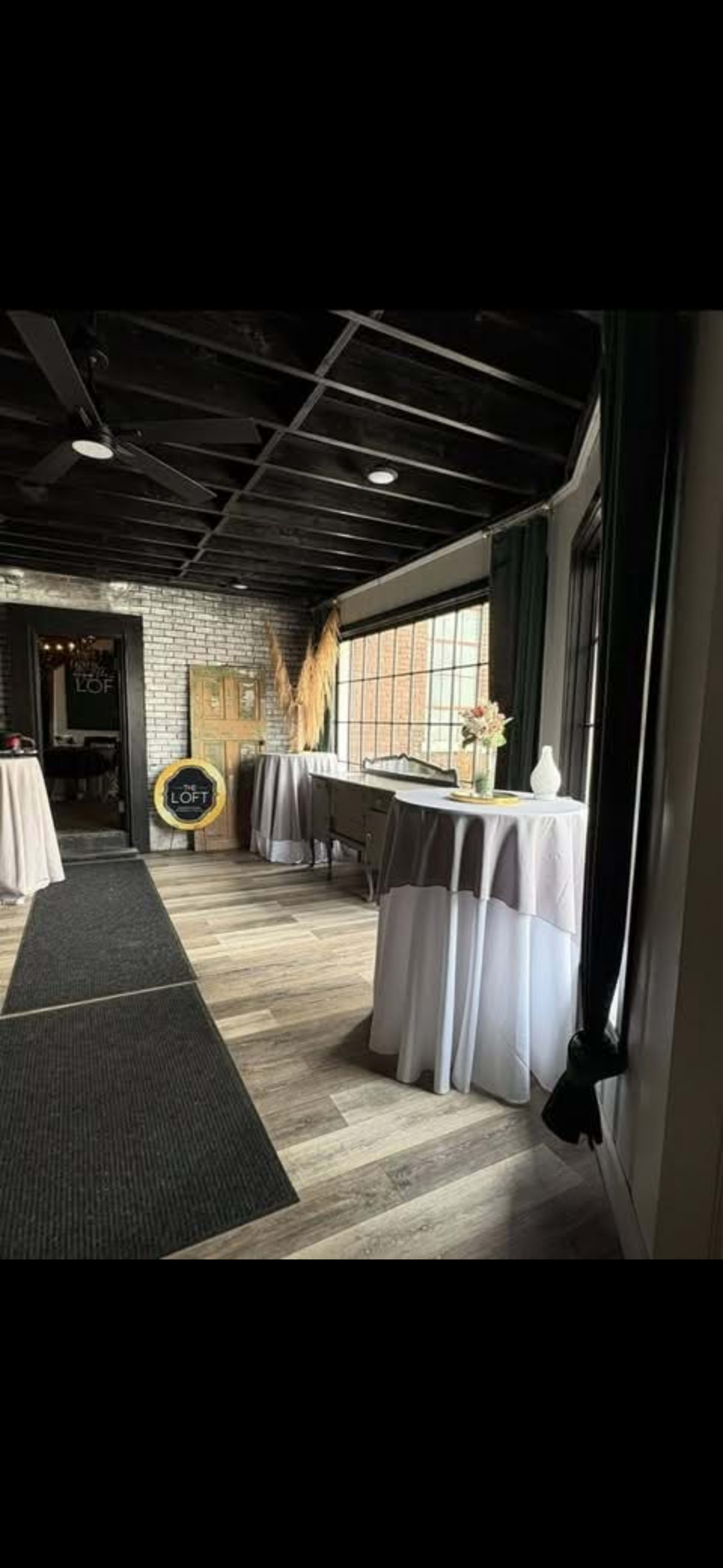 The image shows a spacious interior room with tables covered in white tablecloths, a black mat on the floor, and large windows allowing natural light to enter.