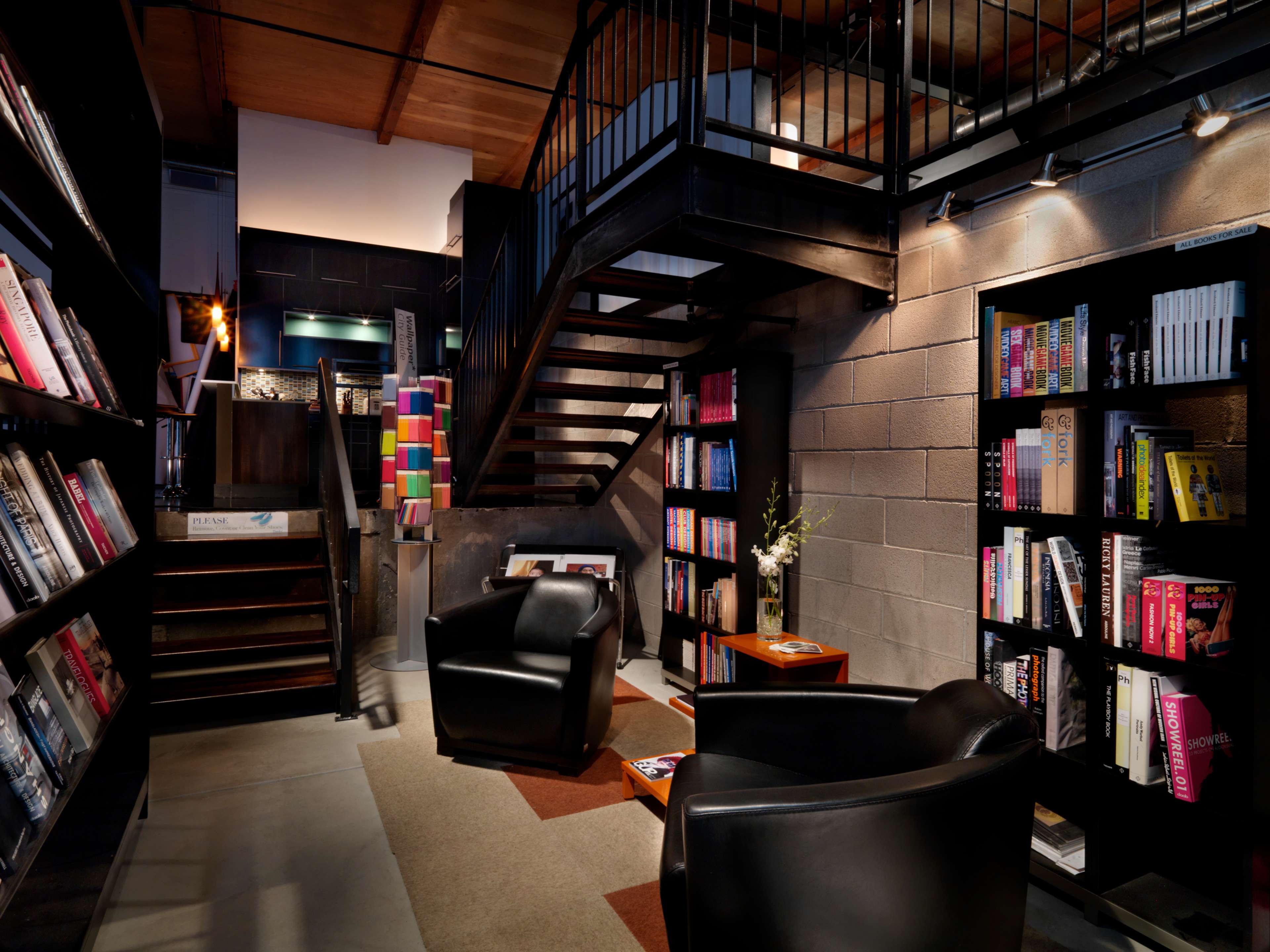 A modern library space with black bookshelves filled with books, a staircase leading to an upper level, and two black chairs facing a wooden side table.