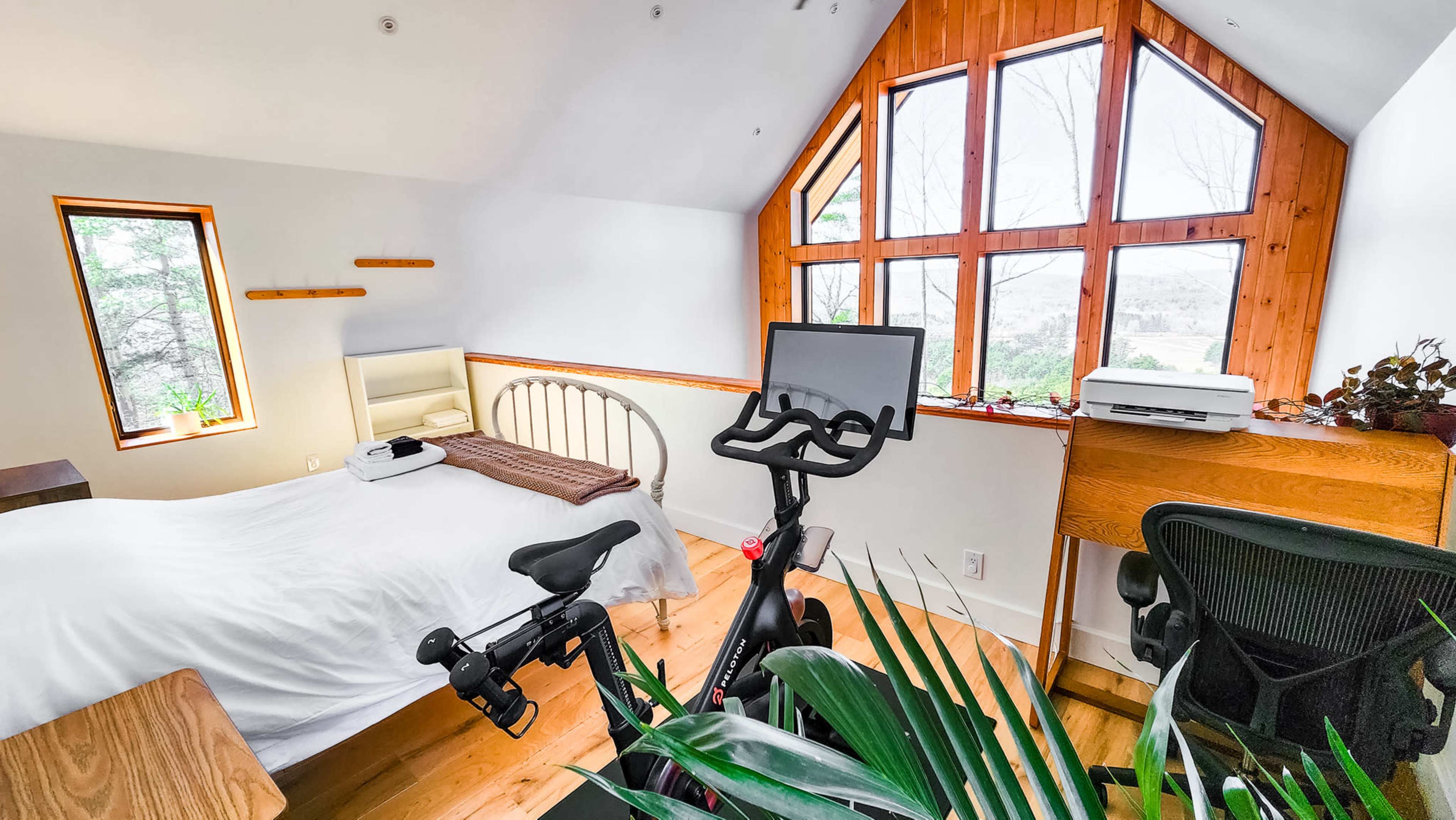 A tidy bedroom with a large window, a bed, a desk, a stationary bike, and a potted plant.