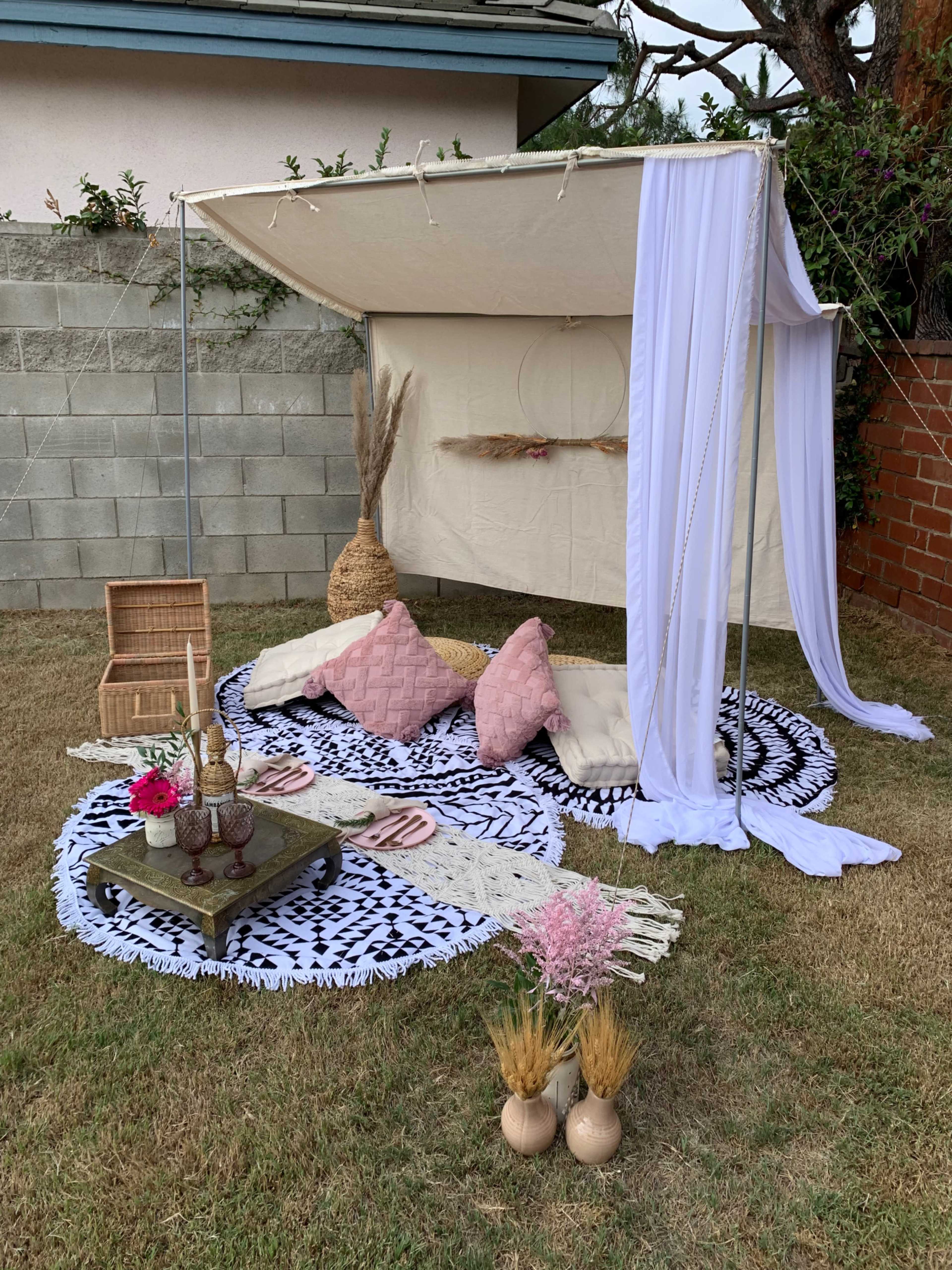 A decorated outdoor seating area with a canopy, featuring cushions, rugs, and decorative items on the grass.