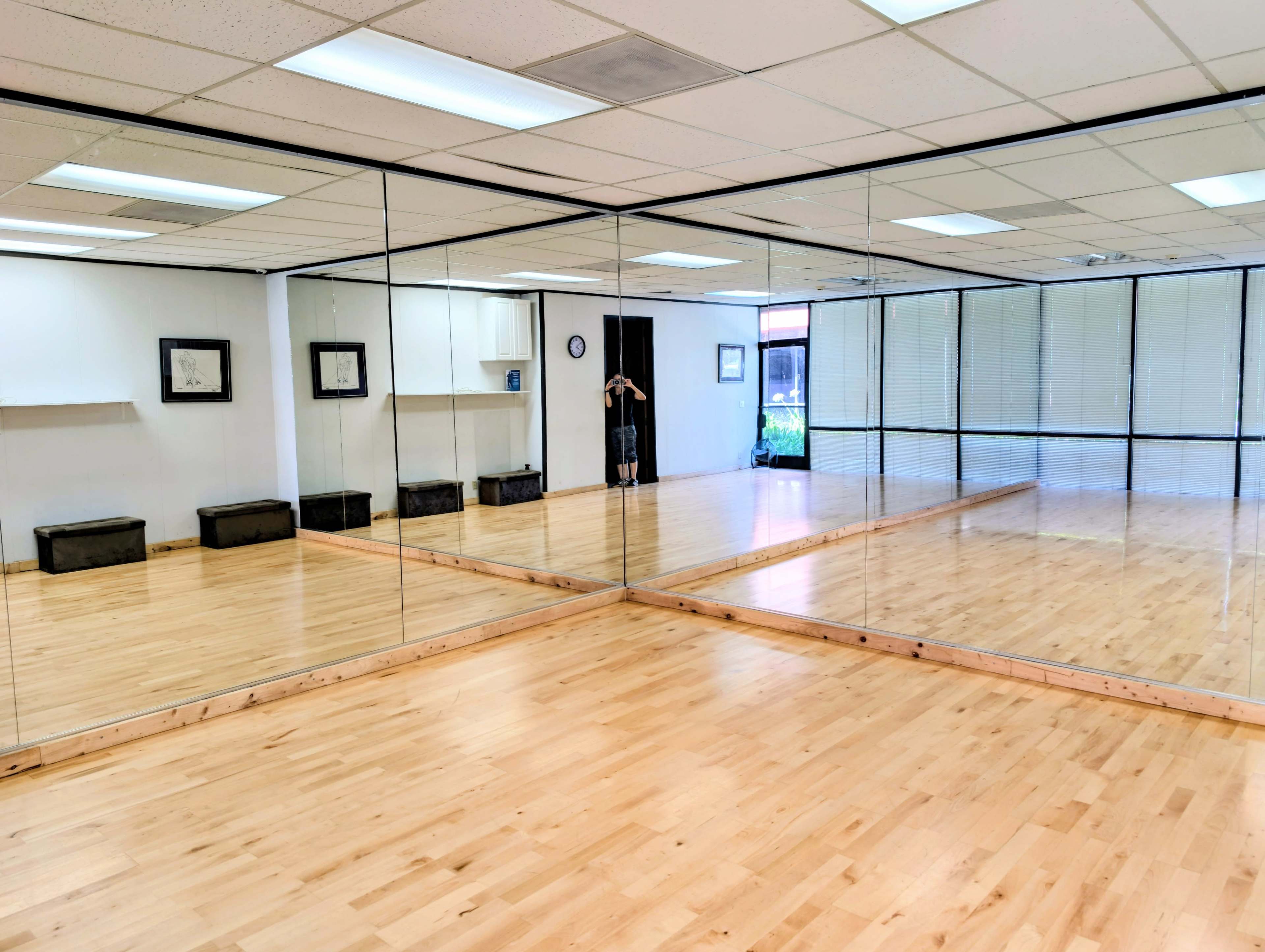 The image shows a brightly lit dance studio with large mirrors on one wall and a wooden floor, featuring shelves and storage boxes along the opposite wall.