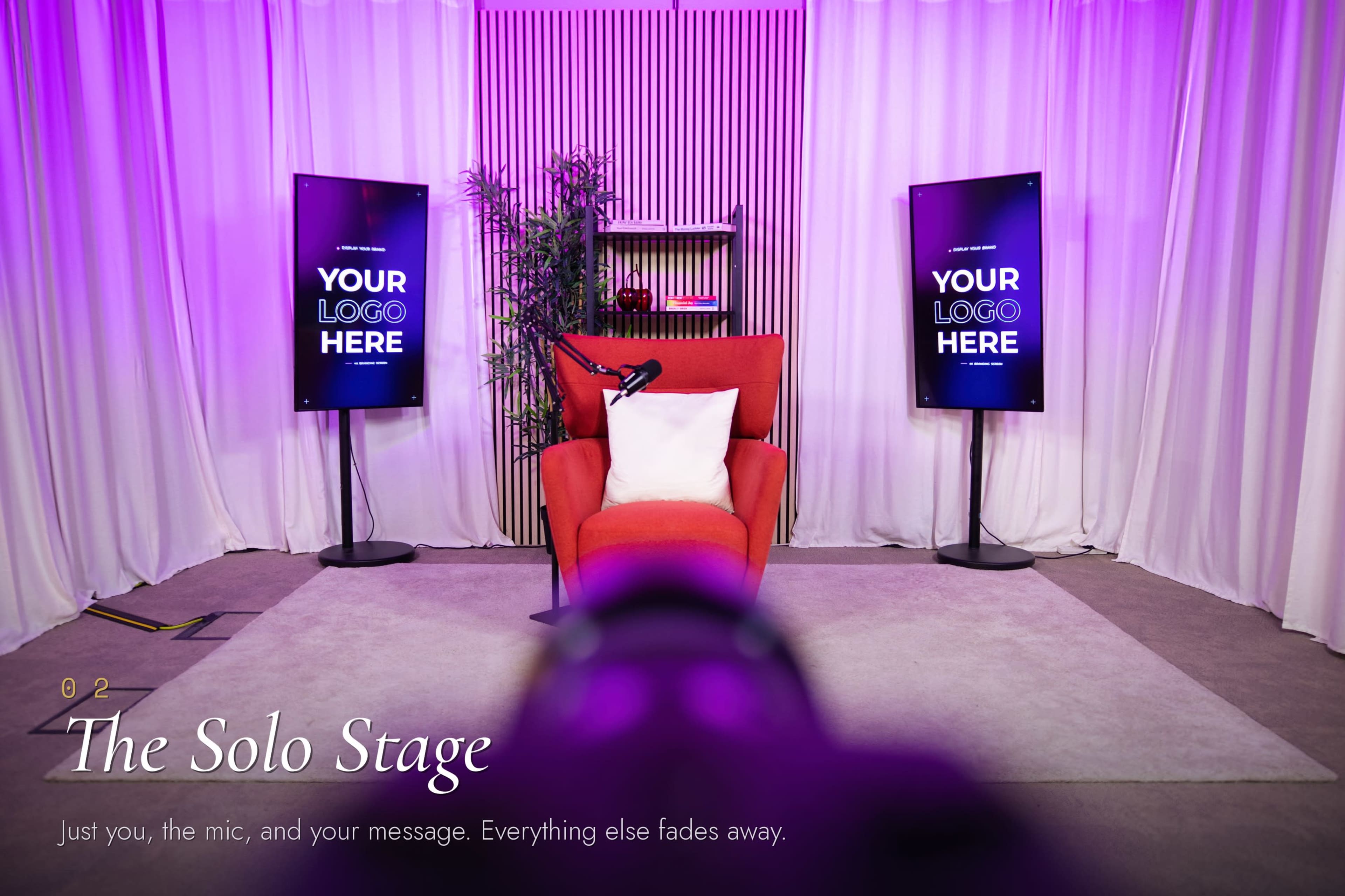 The image shows a stage setup with a red armchair, a microphone, and two screens on either side displaying text that says "YOUR LOGO HERE," all surrounded by purple lighting and white curtains.