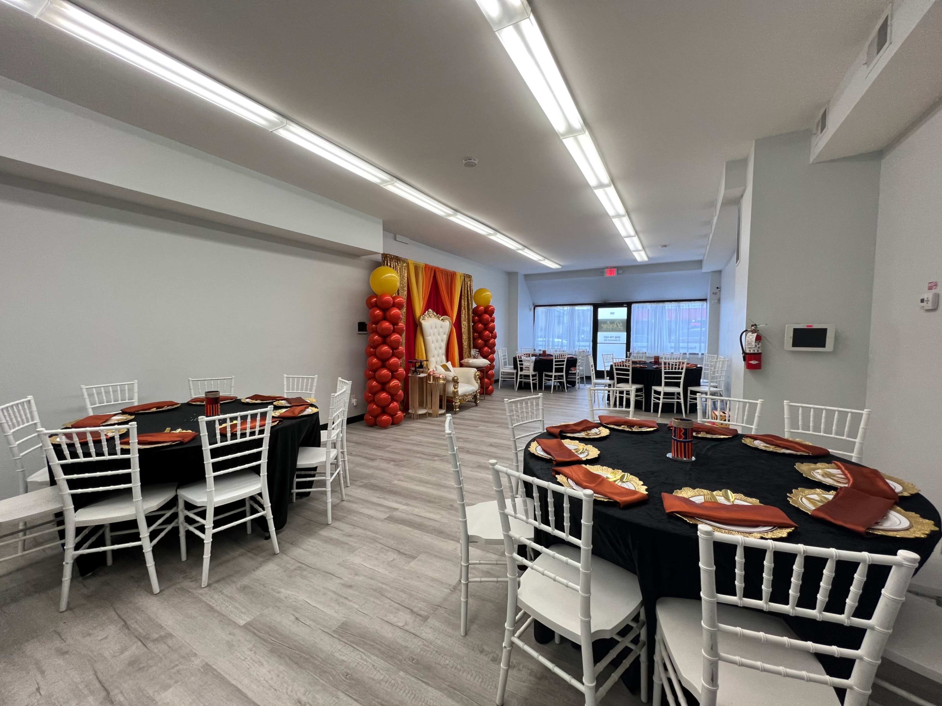 The image shows a banquet room with several round tables covered in black tablecloths and adorned with gold accents, along with a decorative backdrop featuring red and yellow balloons.