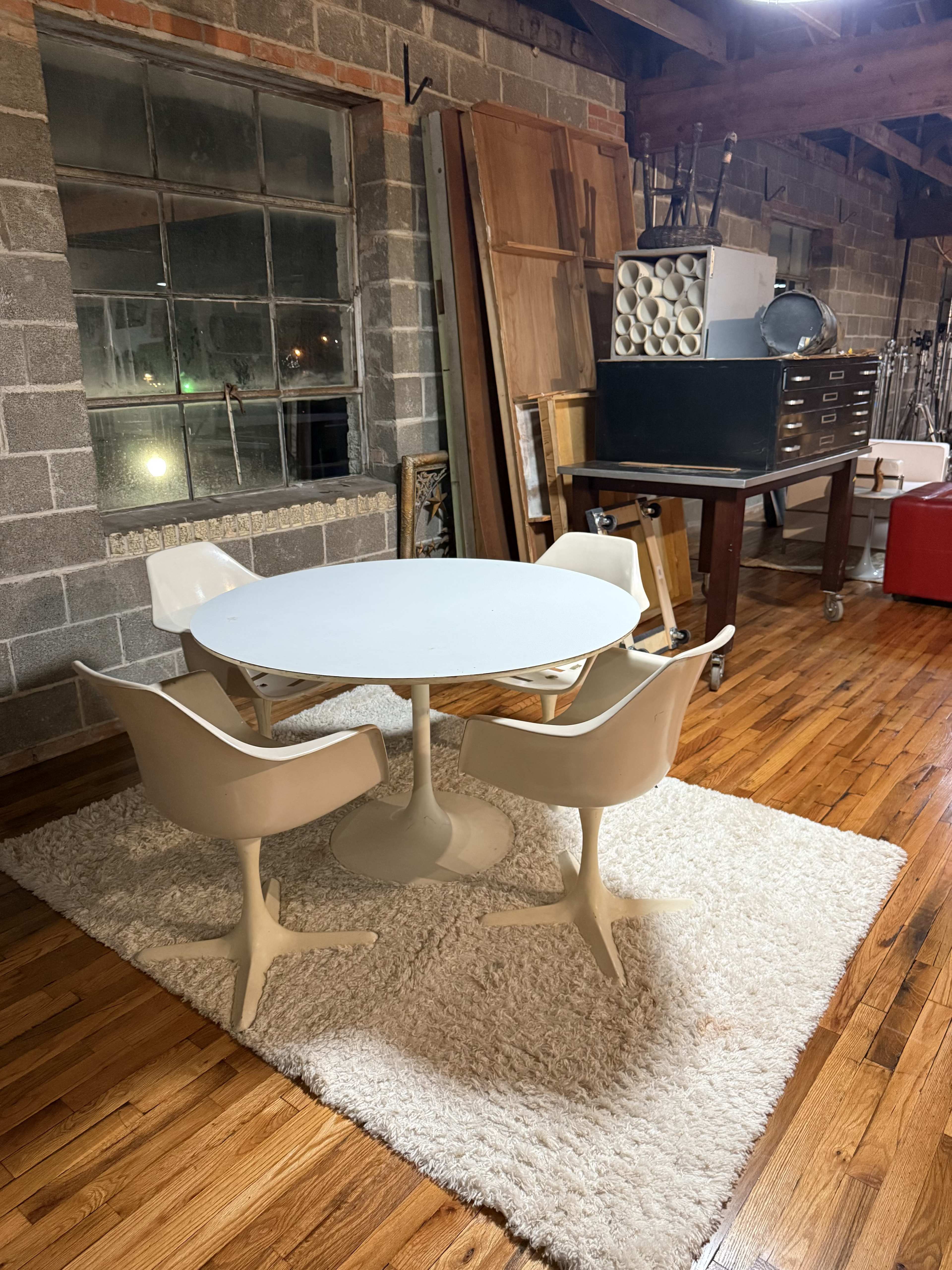 The image shows a minimalist dining area with a round white table and three matching chairs on a shaggy white rug, situated against a brick wall with large windows.