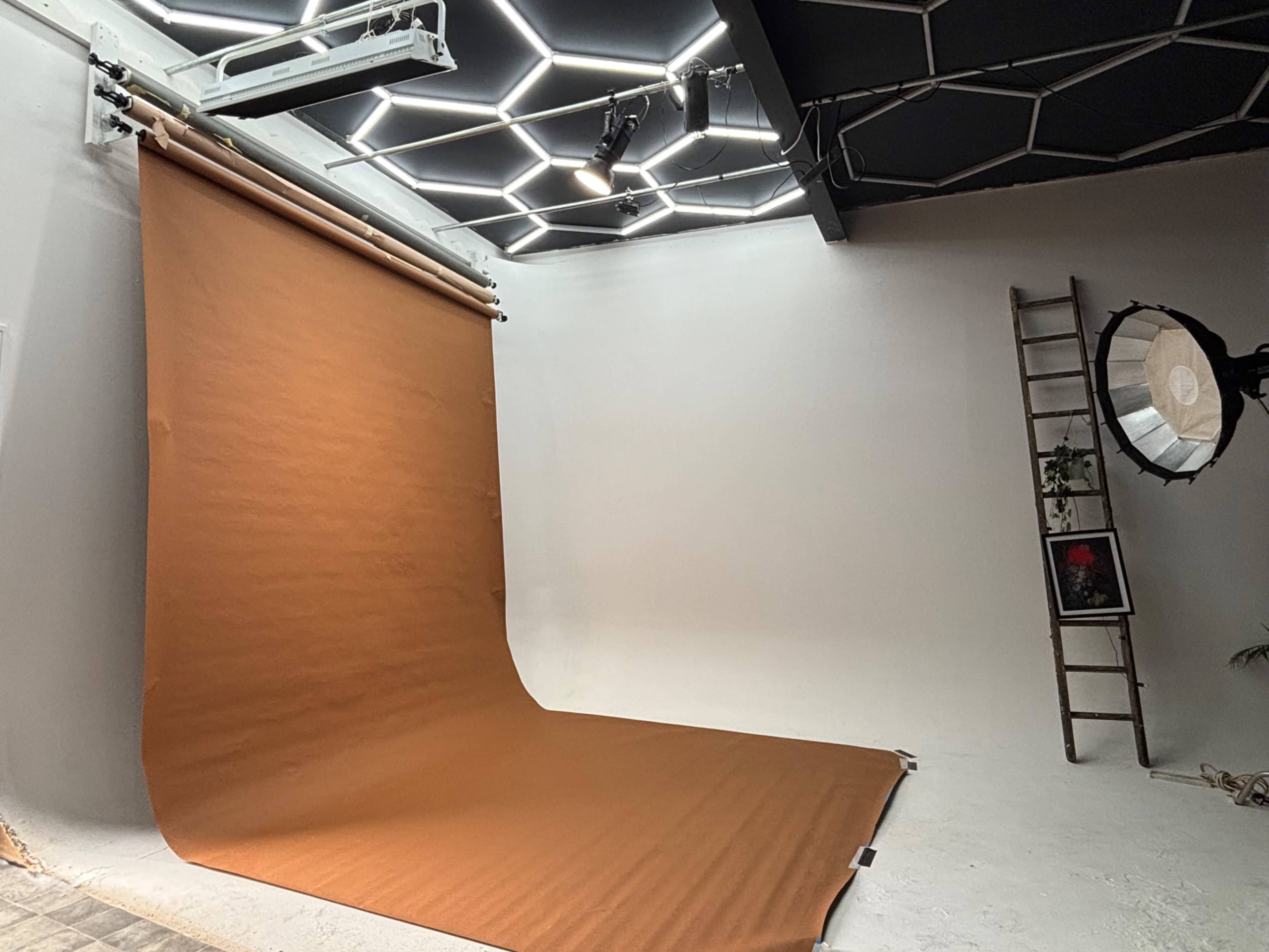 The image shows a photography studio with a brown backdrop, a ladder, and a softbox lighting setup under a geometric ceiling.