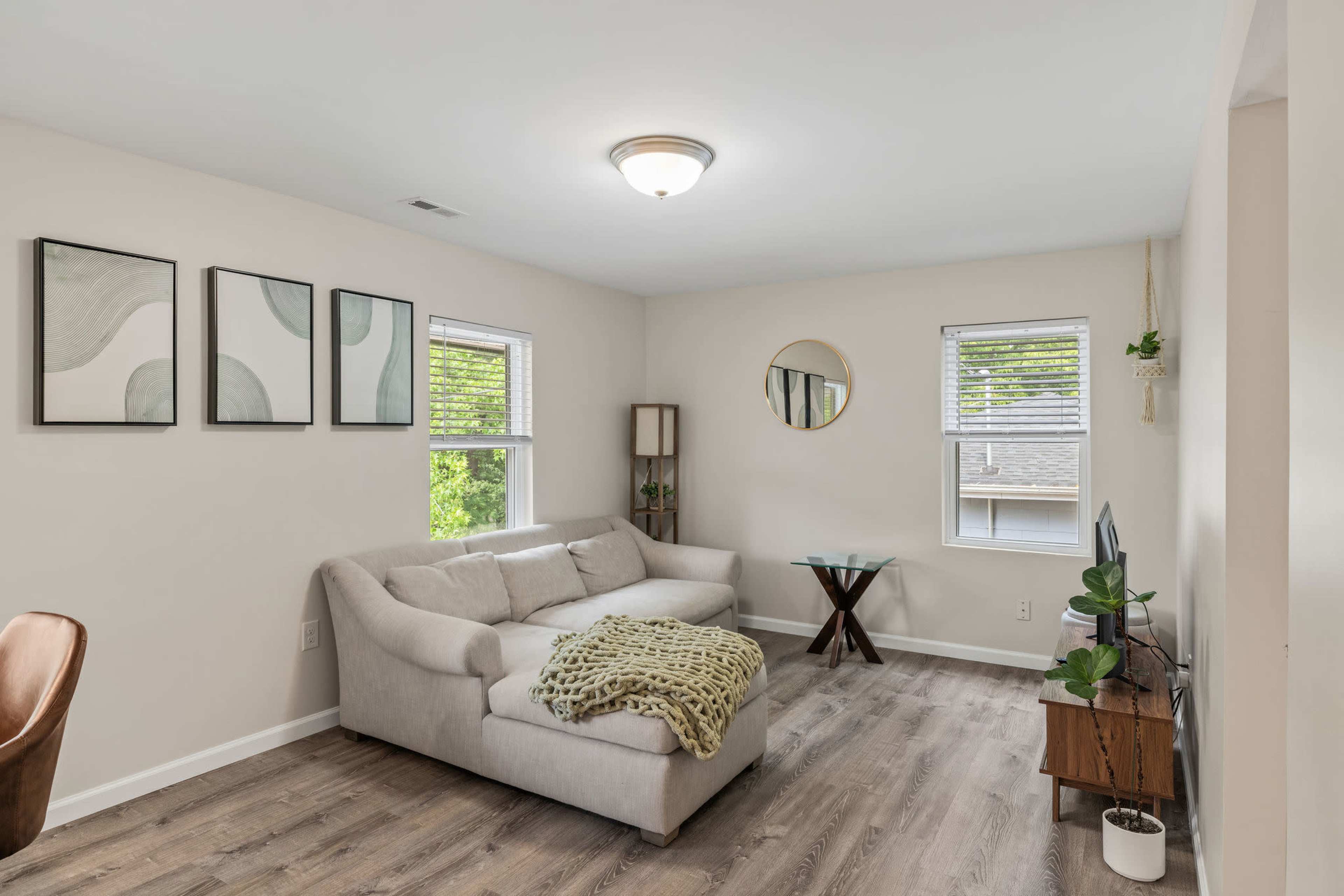 The living room features a light-colored couch, a small table, and framed artwork on the walls, with two windows providing natural light.