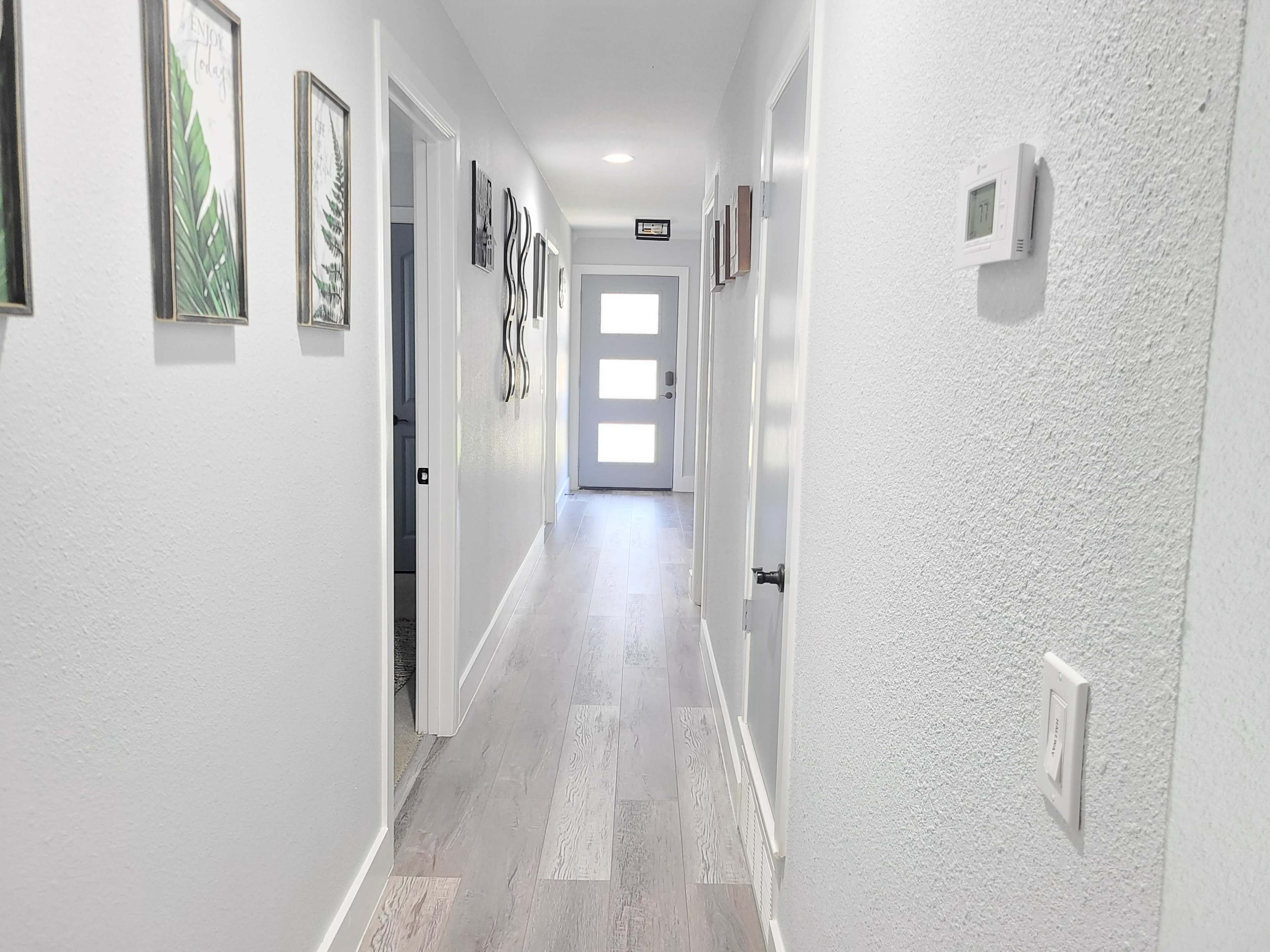A long hallway features light-colored walls, wooden flooring, and several framed artworks, leading to a door with a glass panel at the end.