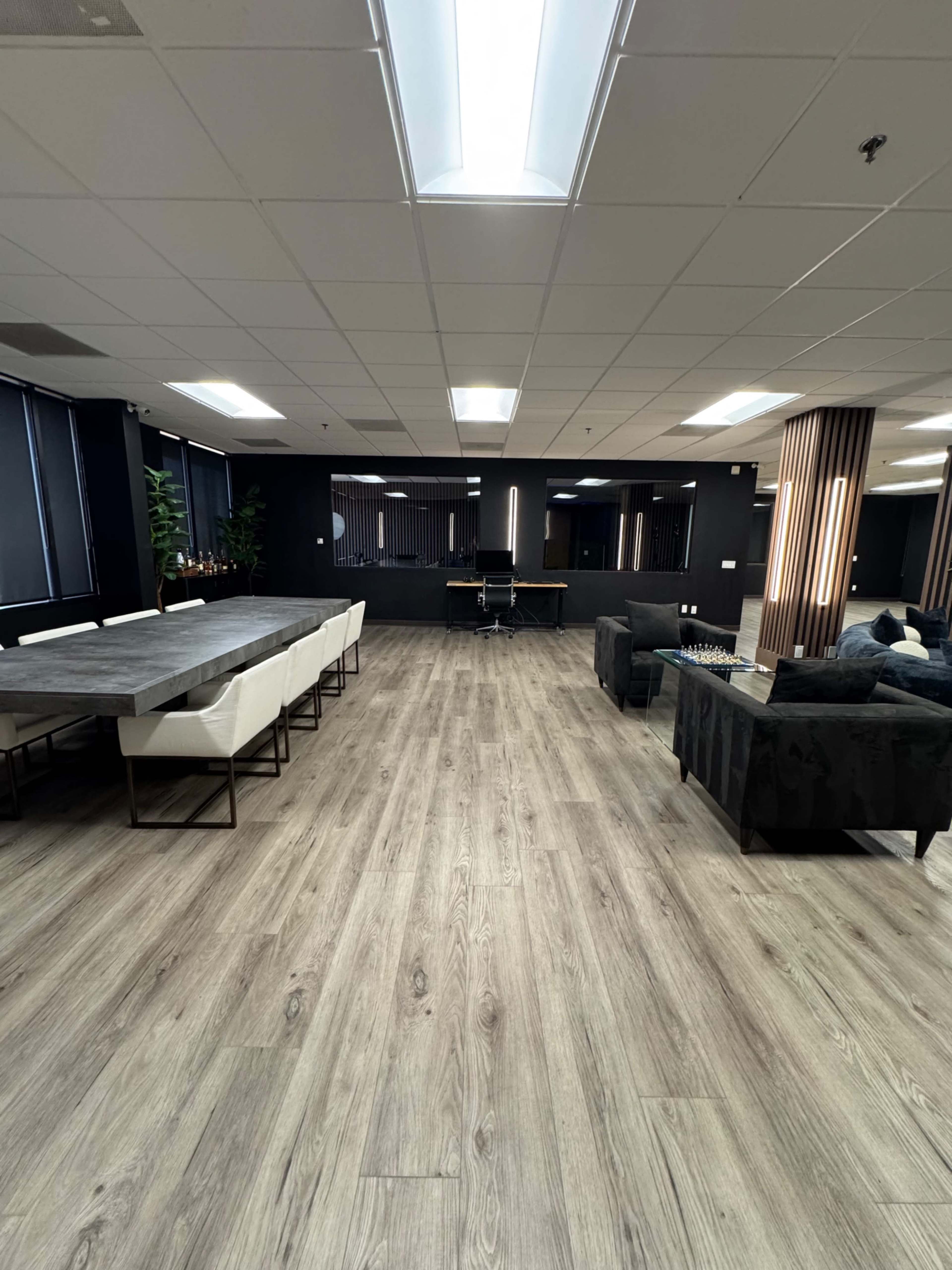 The image shows a spacious office interior featuring a long dining table with chairs, a lounge area with sofas, and a large wooden floor.