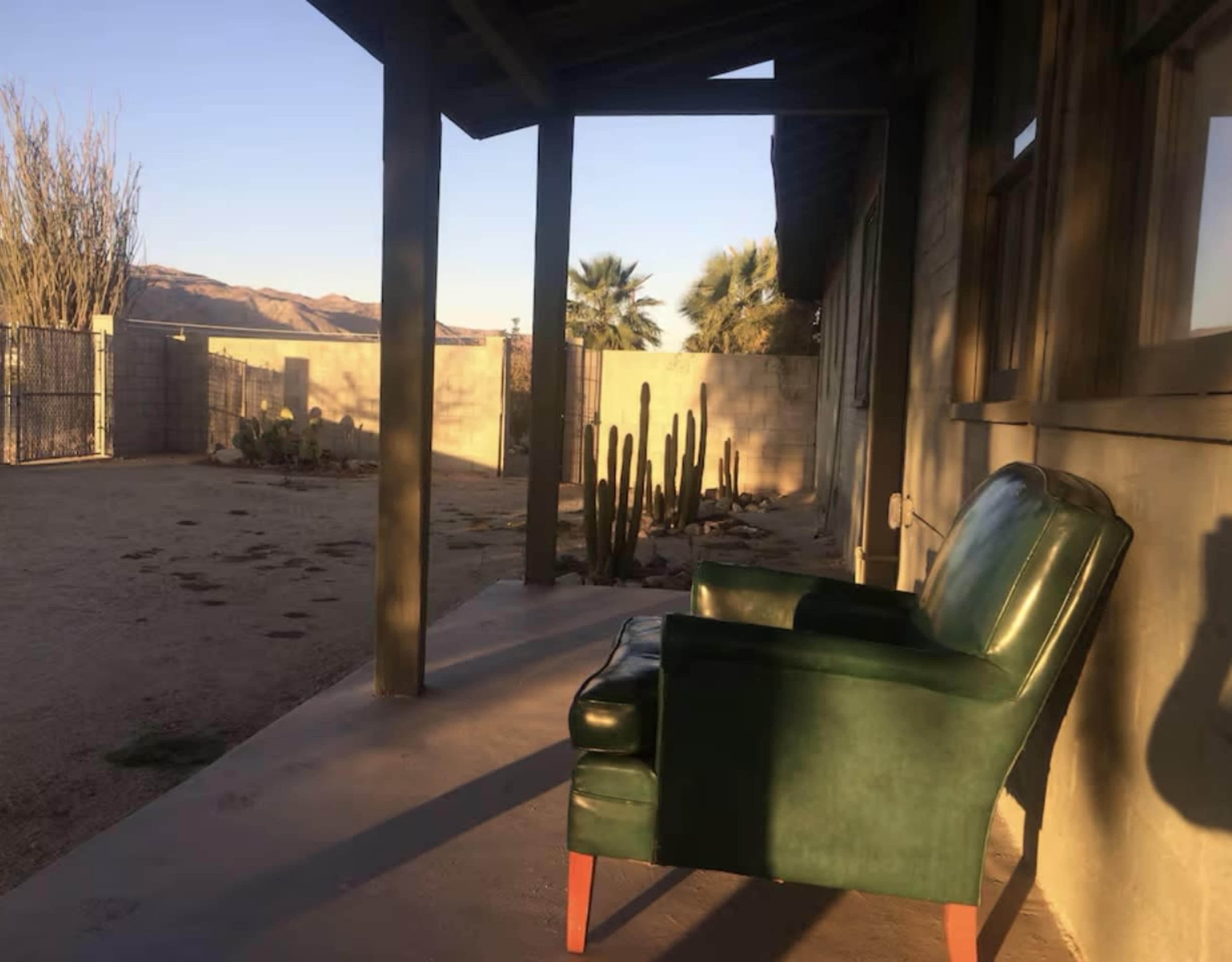 A green chair sits on a concrete porch in front of a desert landscape featuring cacti and distant hills.