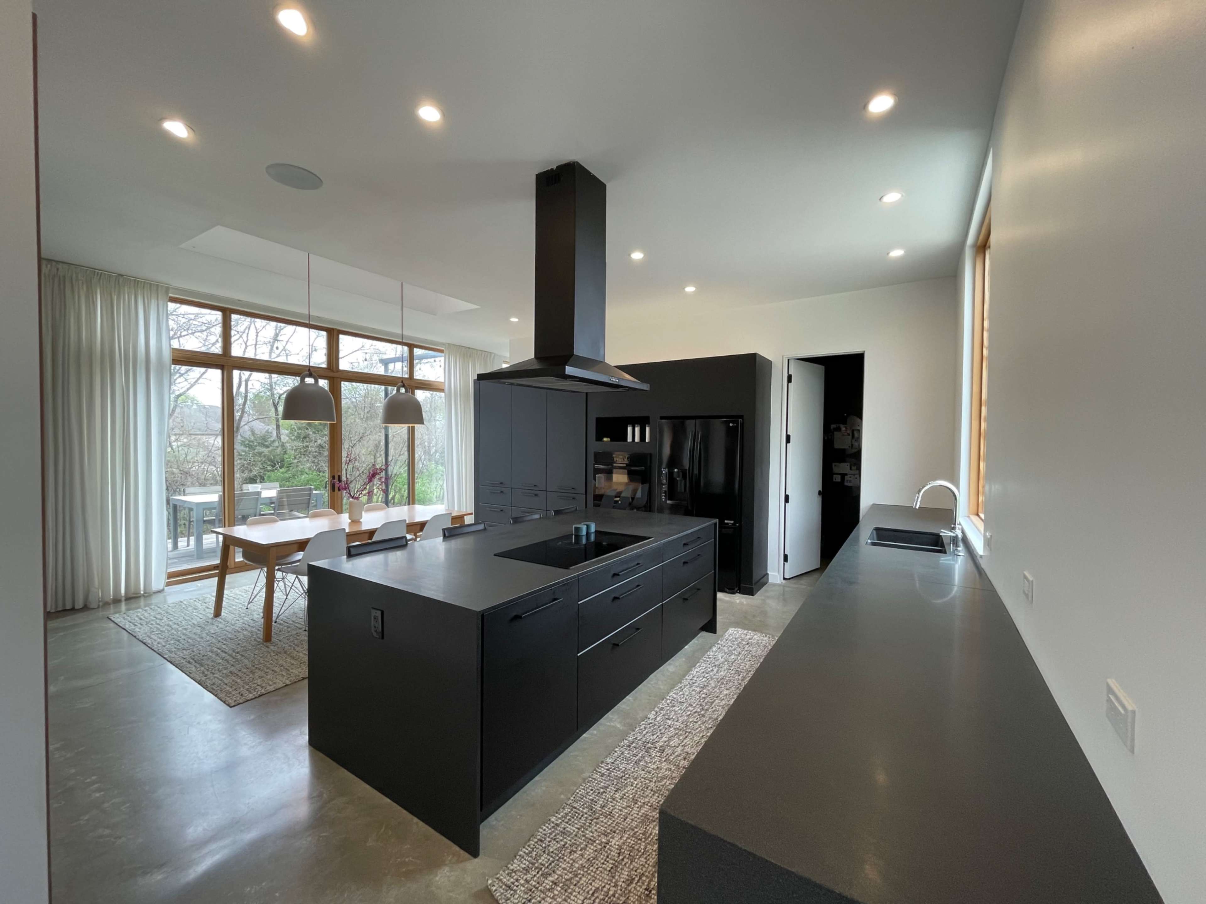 A modern kitchen features a large black island with an integrated sink, sleek cabinetry, and a ventilation hood, with natural light streaming through large windows.