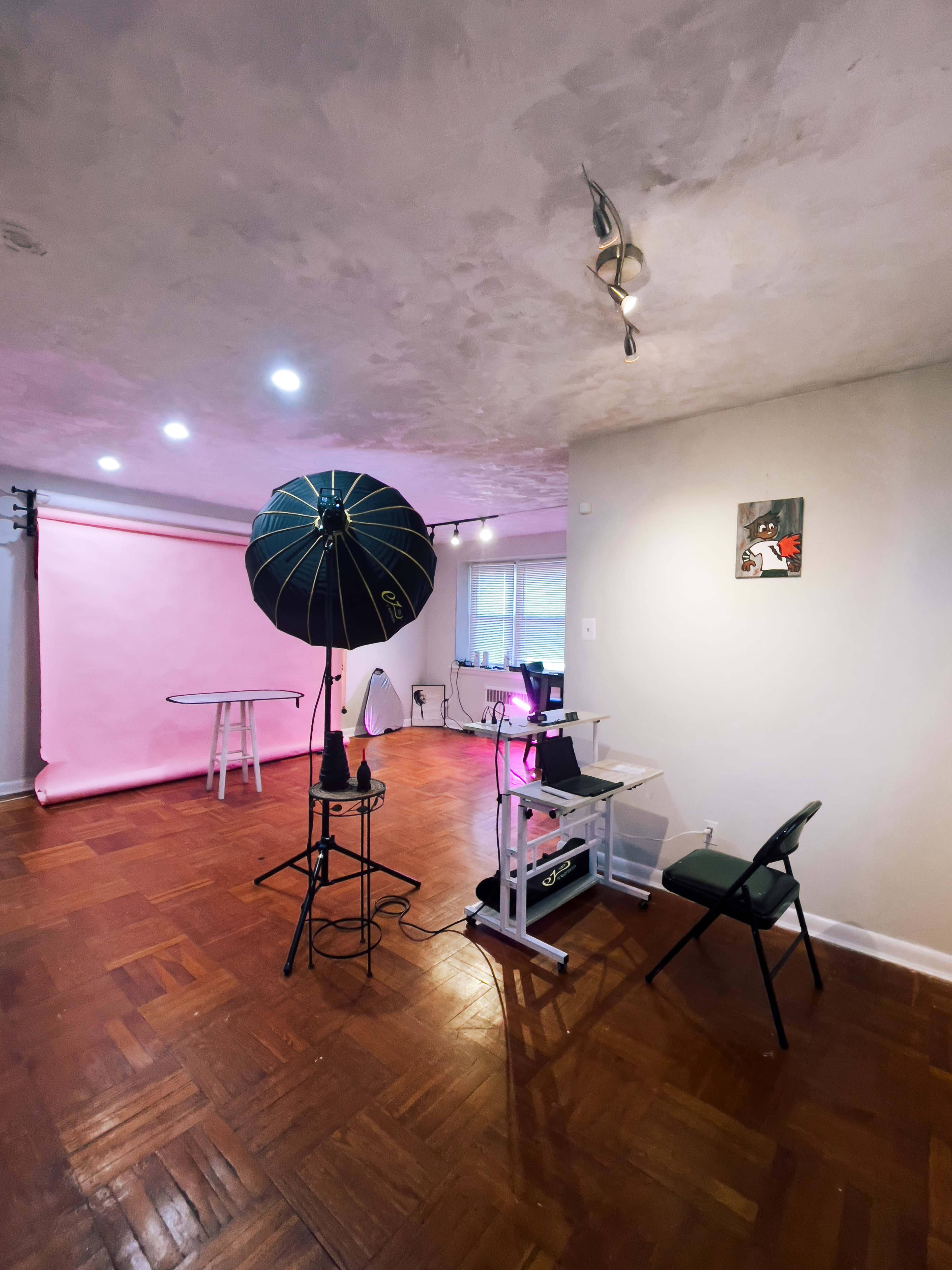 A bright studio space features a pink backdrop, a photography umbrella light, and a small desk setup.
