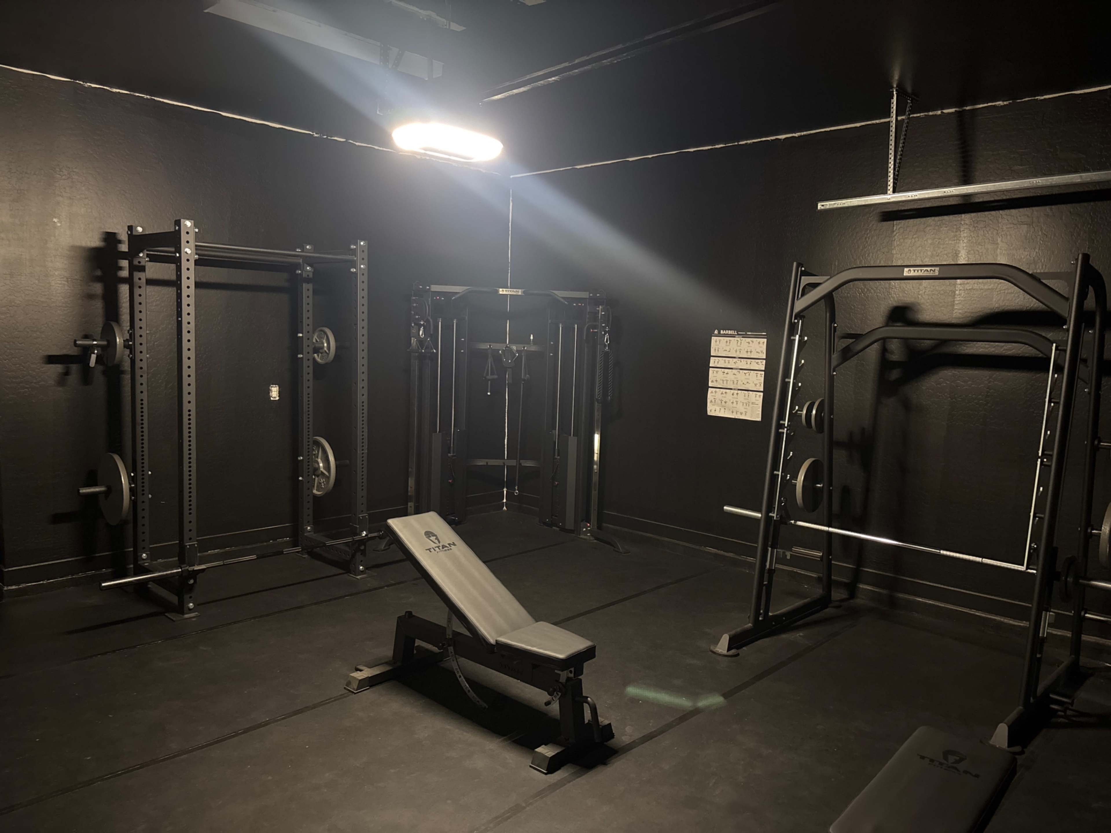 The image shows a dimly lit gym space equipped with various strength training machines and a bench.