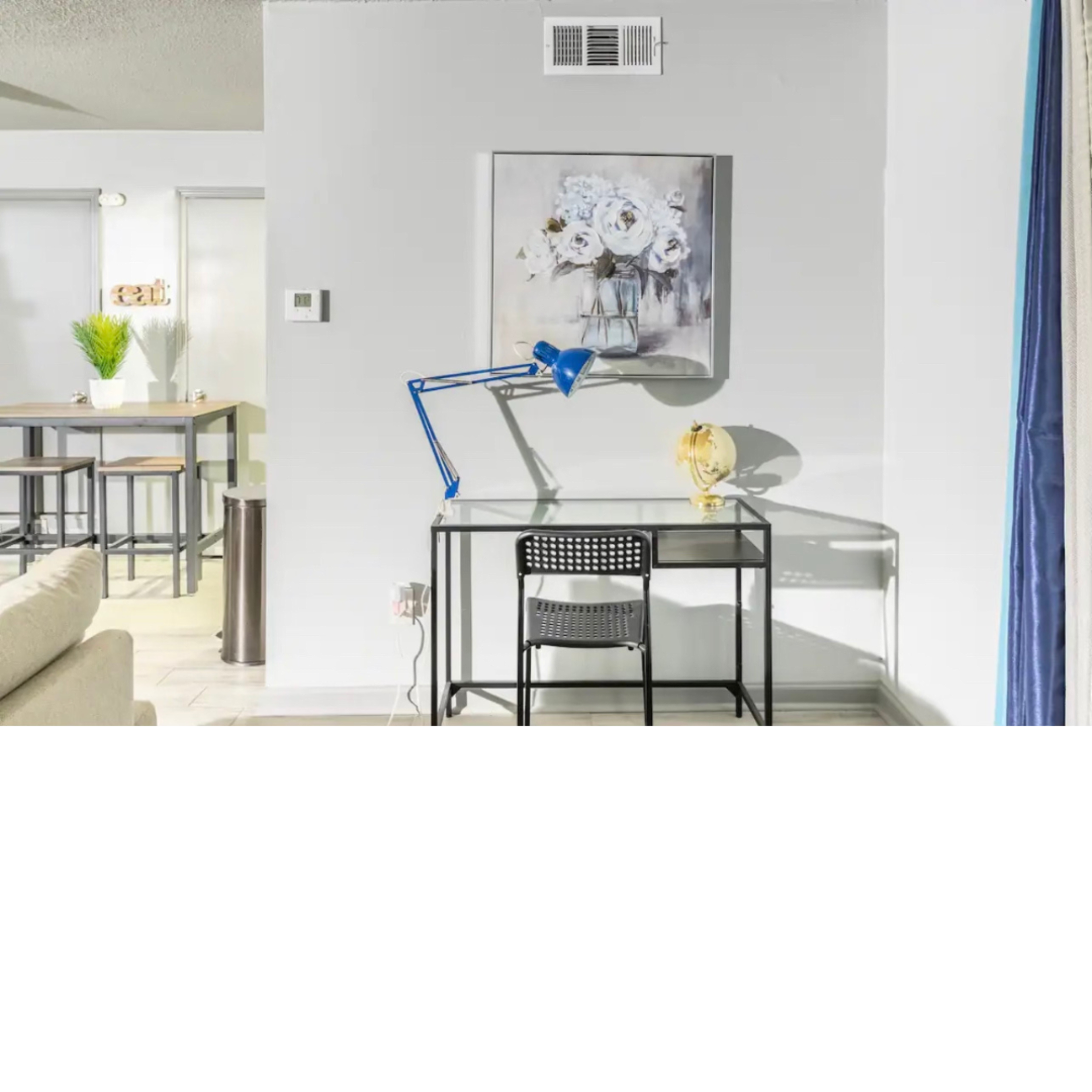 A small desk with a blue lamp, a chair, and a floral painting on the wall beside a light-filled room with a sofa and a dining area in the background.