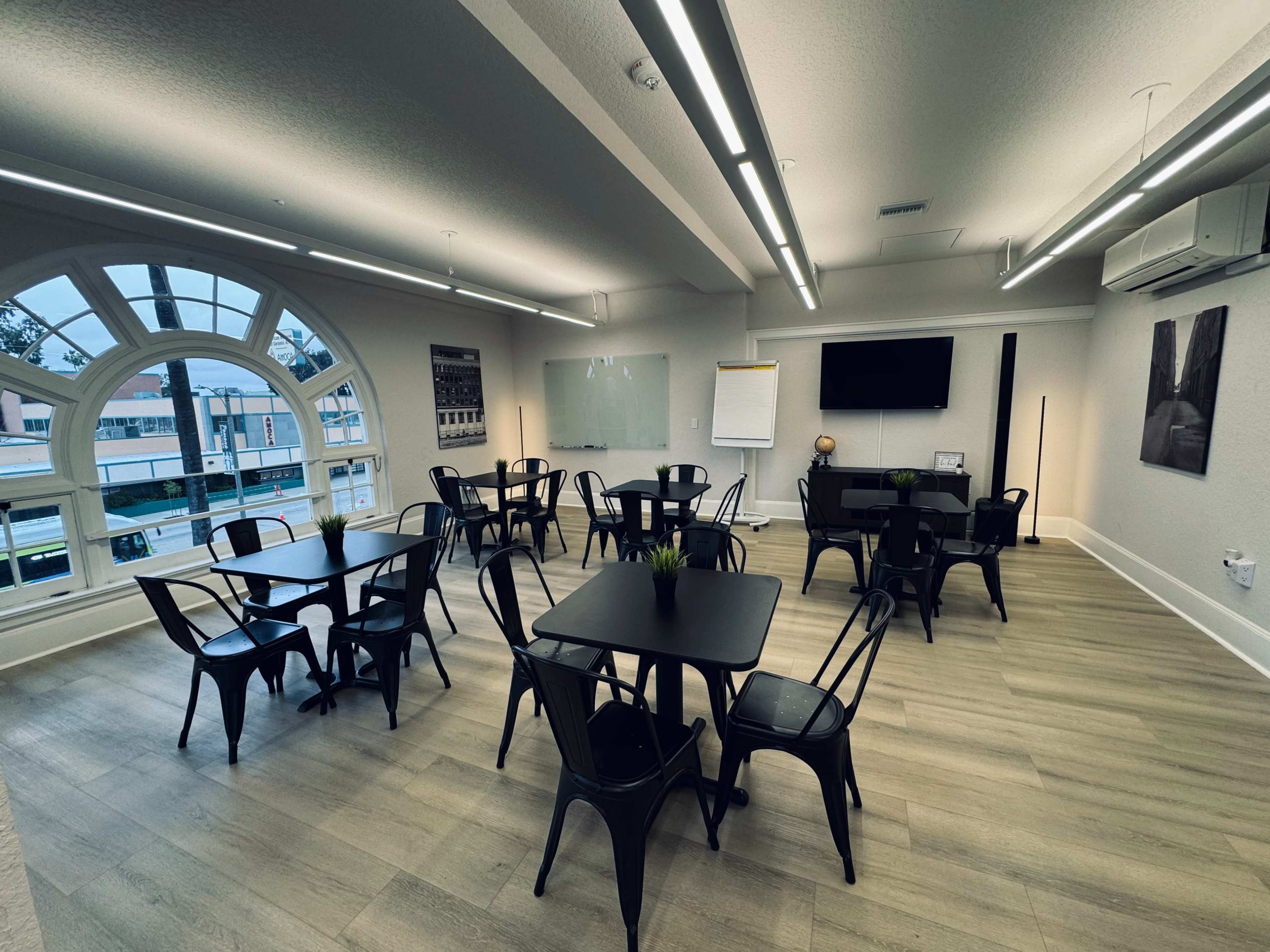 A meeting room features several black tables and chairs, large windows, and a presentation screen on the wall.