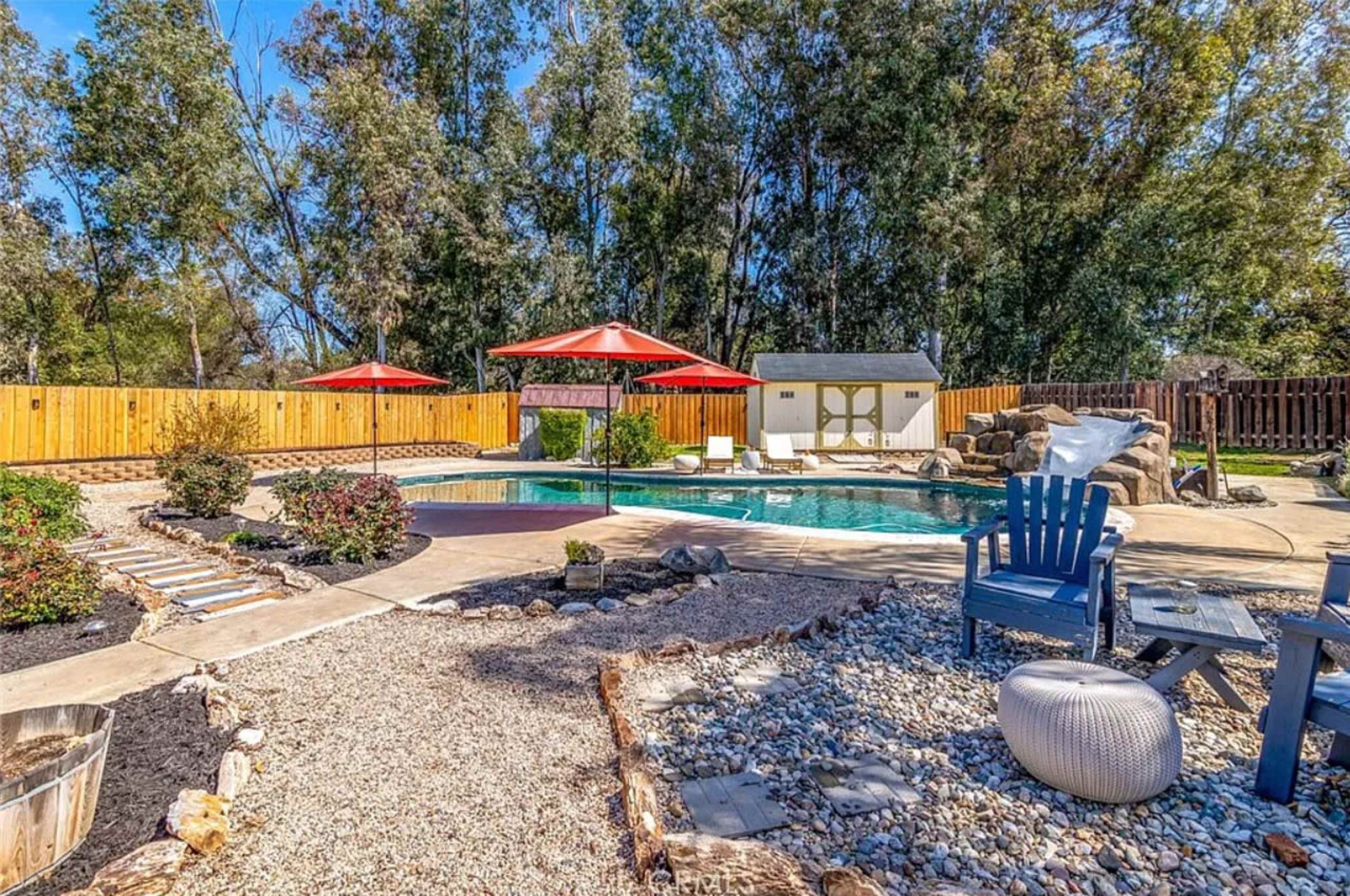 A backyard with a swimming pool, patio seating under red umbrellas, landscaped greenery, and gravel paths.