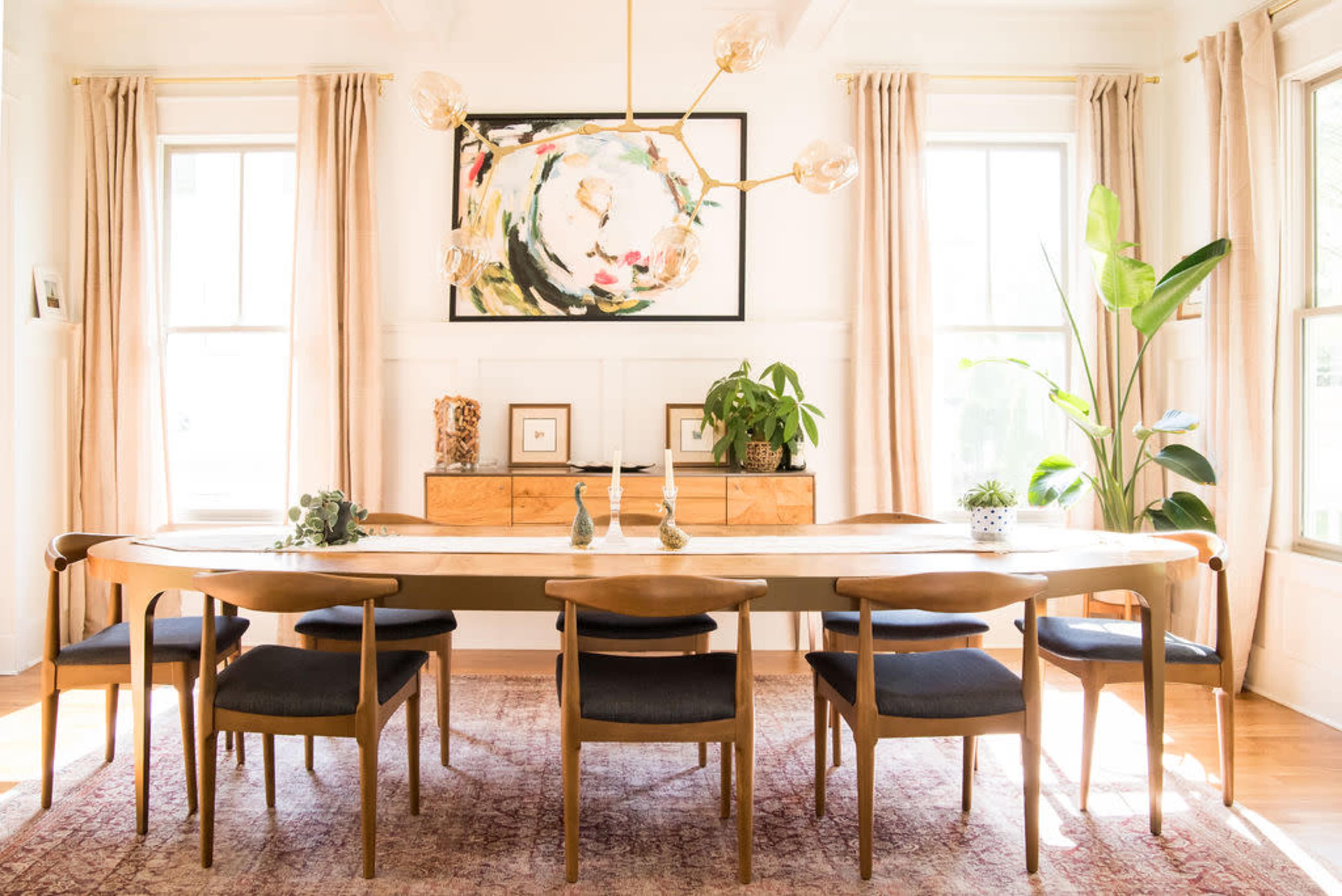 A modern dining room features a long wooden table surrounded by chairs, with large windows, artwork on the wall, and plants in the corners.