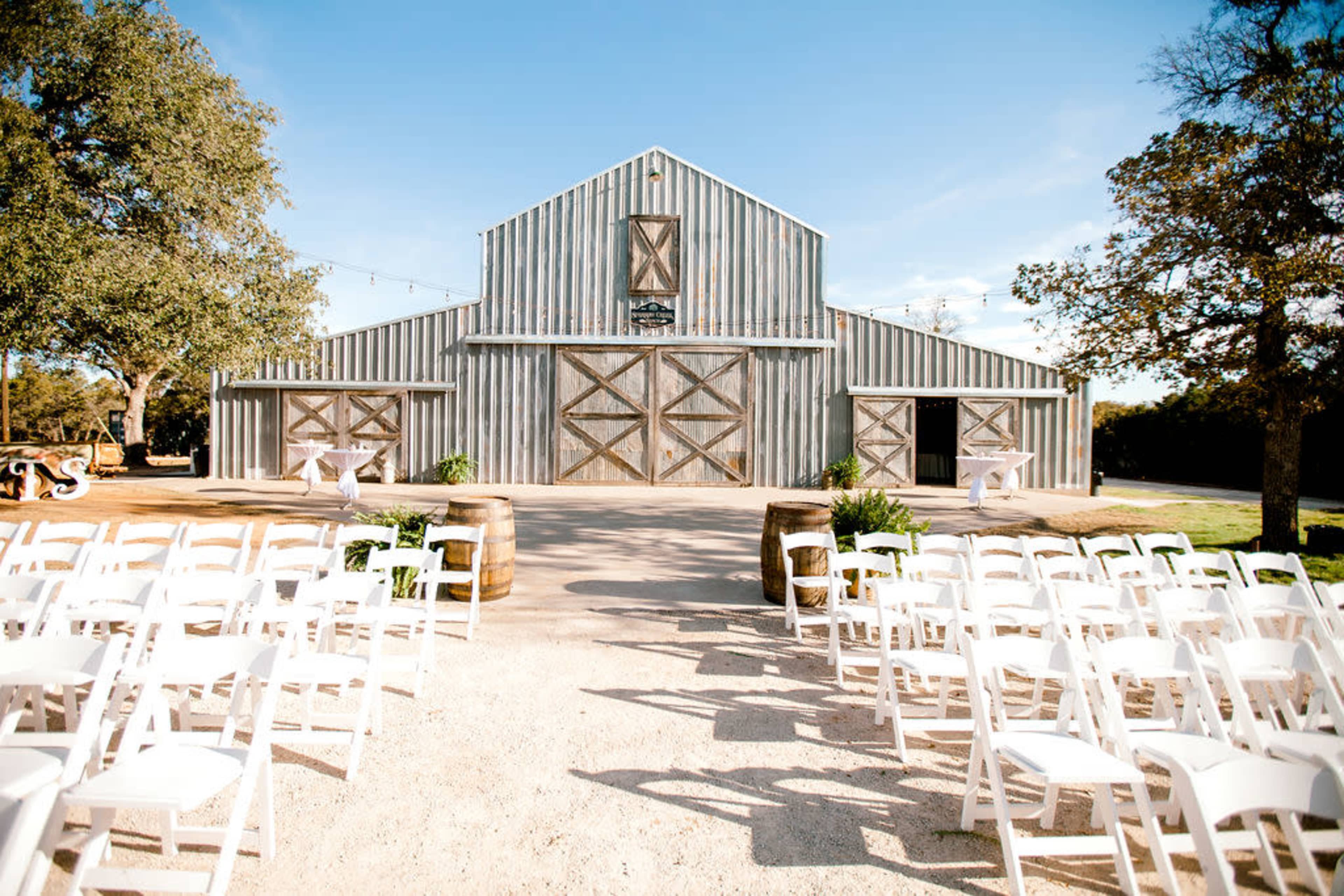 The image shows a rustic barn with a gravel path leading to it, surrounded by white folding chairs arranged for an outdoor event.